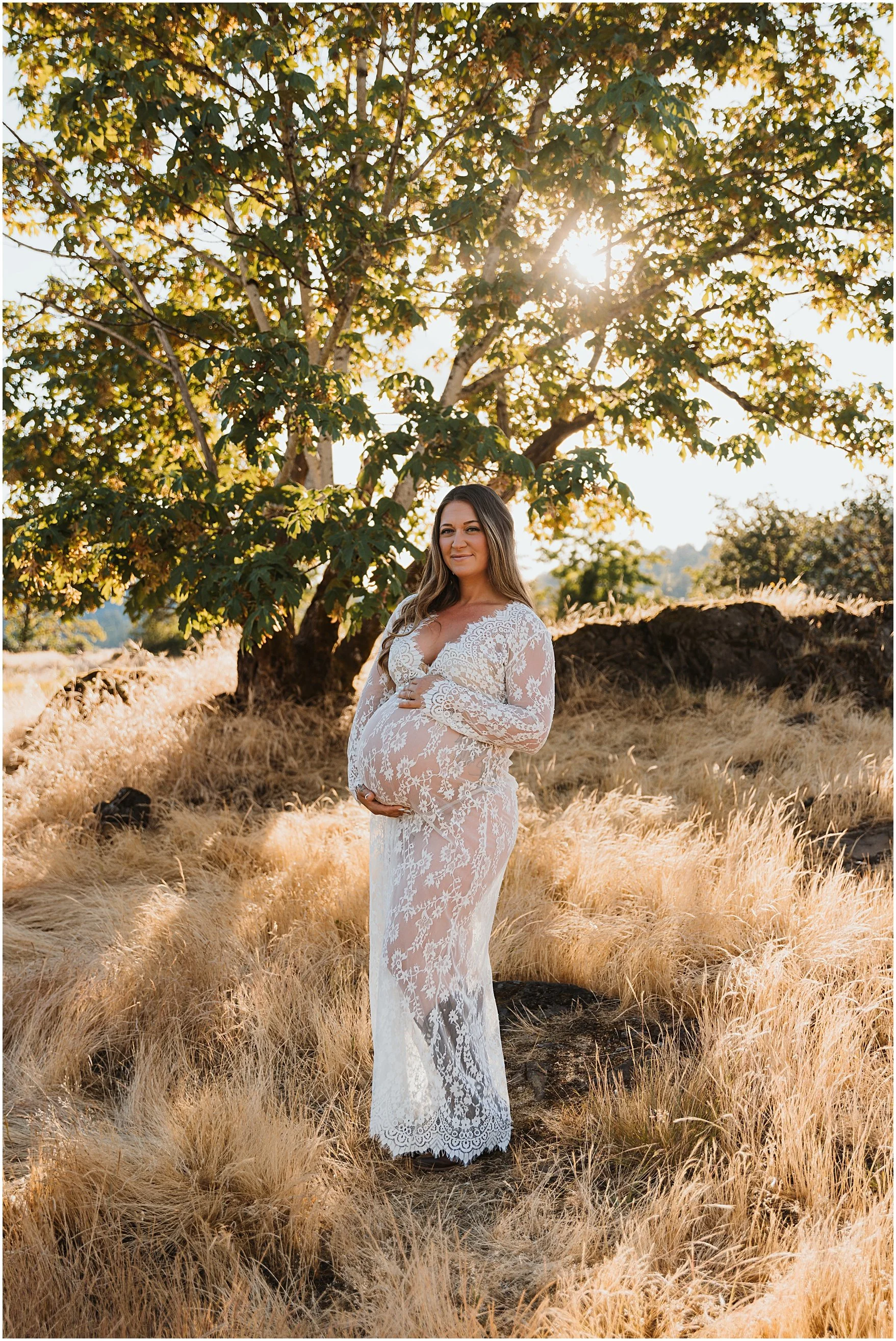 A pregnant woman wearing a white lace dress standing outdoors in a field of tall, golden grass, with a large leafy tree behind her and the sun shining through the branches.