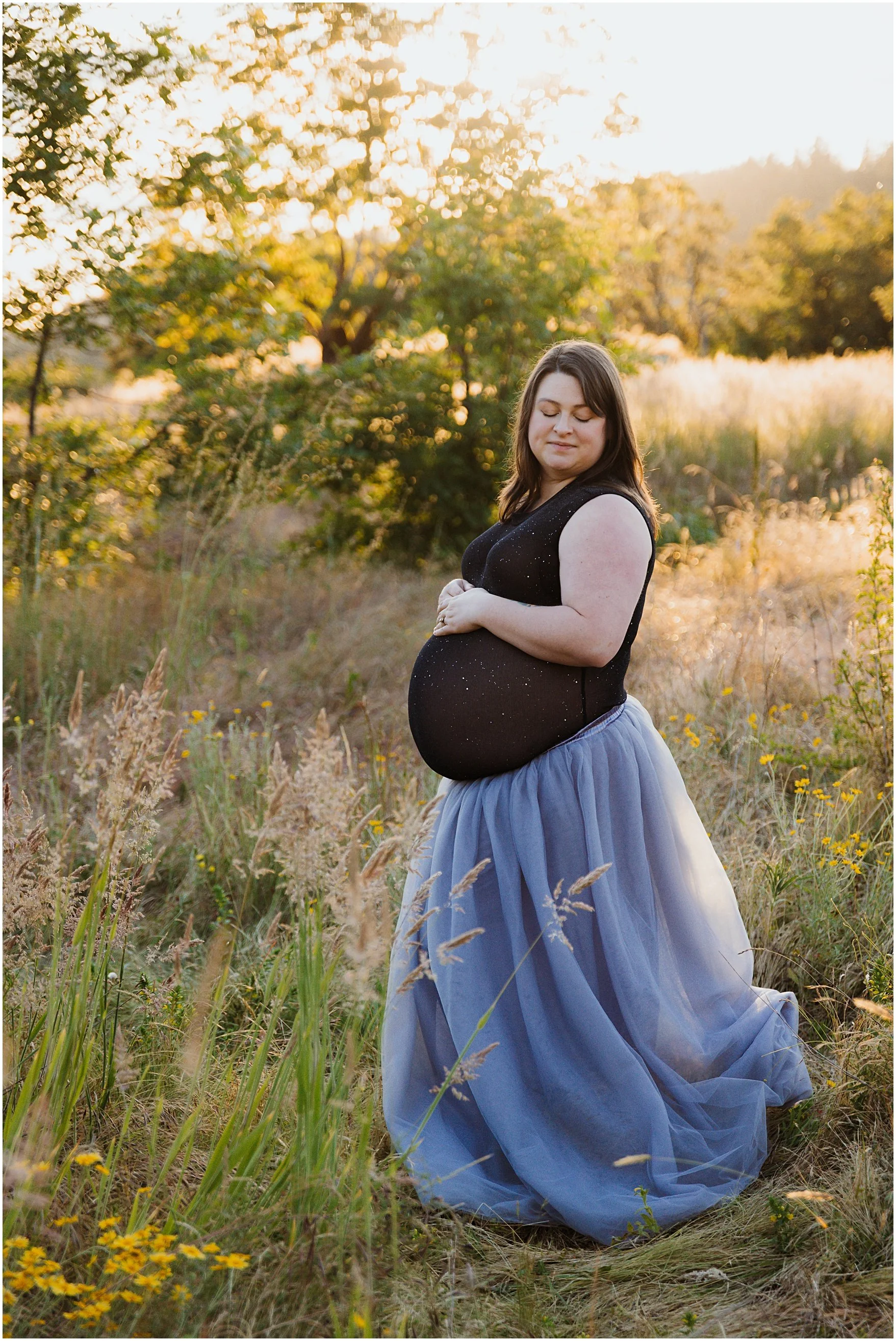 A pregnant woman kneeling in a field at sunset, wearing a black top and a blue tulle skirt, with her hands resting on her belly and eyes closed.