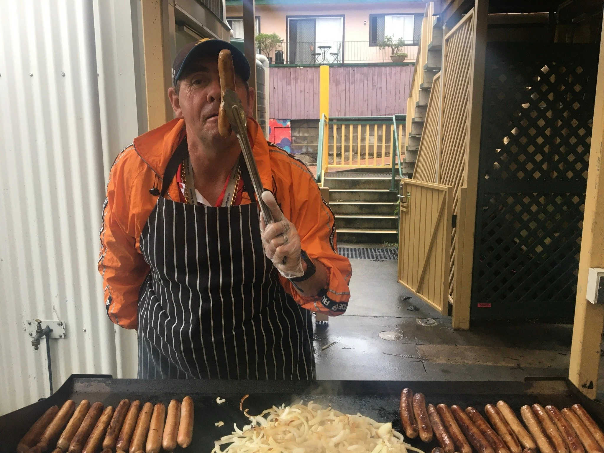 A man wearing an apron holds a sausage above a barbeque of sausages and onion.
