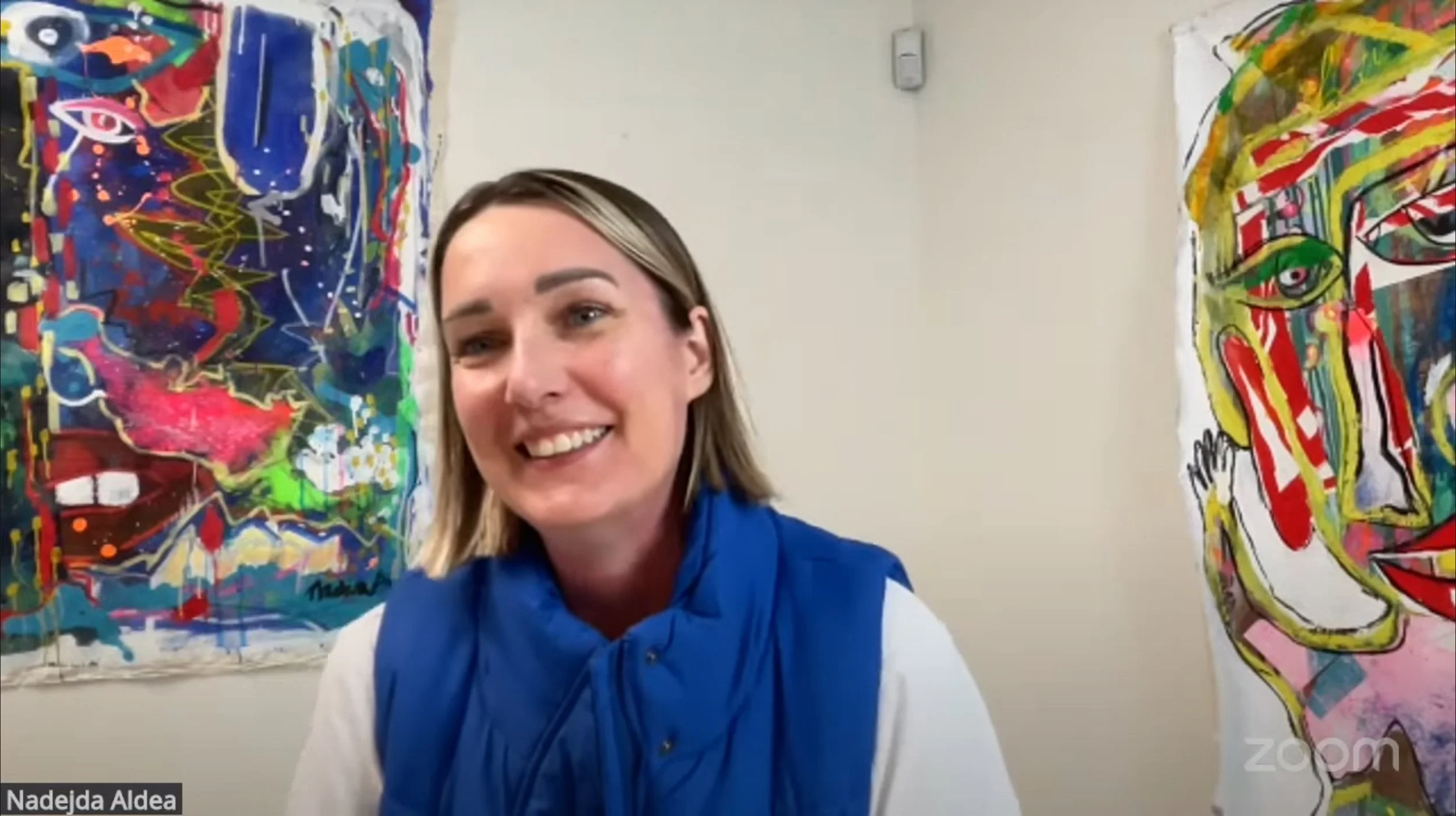 Smiling woman with blonde hair wearing a blue vest and white shirt, sitting in front of colorful abstract paintings on a plain wall.