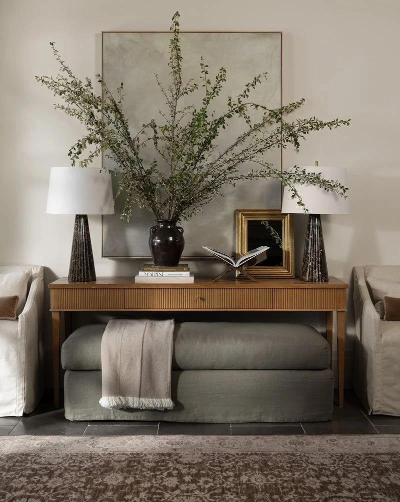 Elegant console table with tall greenery in a dark vase, modern lamps, layered artwork, and a neutral bench for a refined living room look.