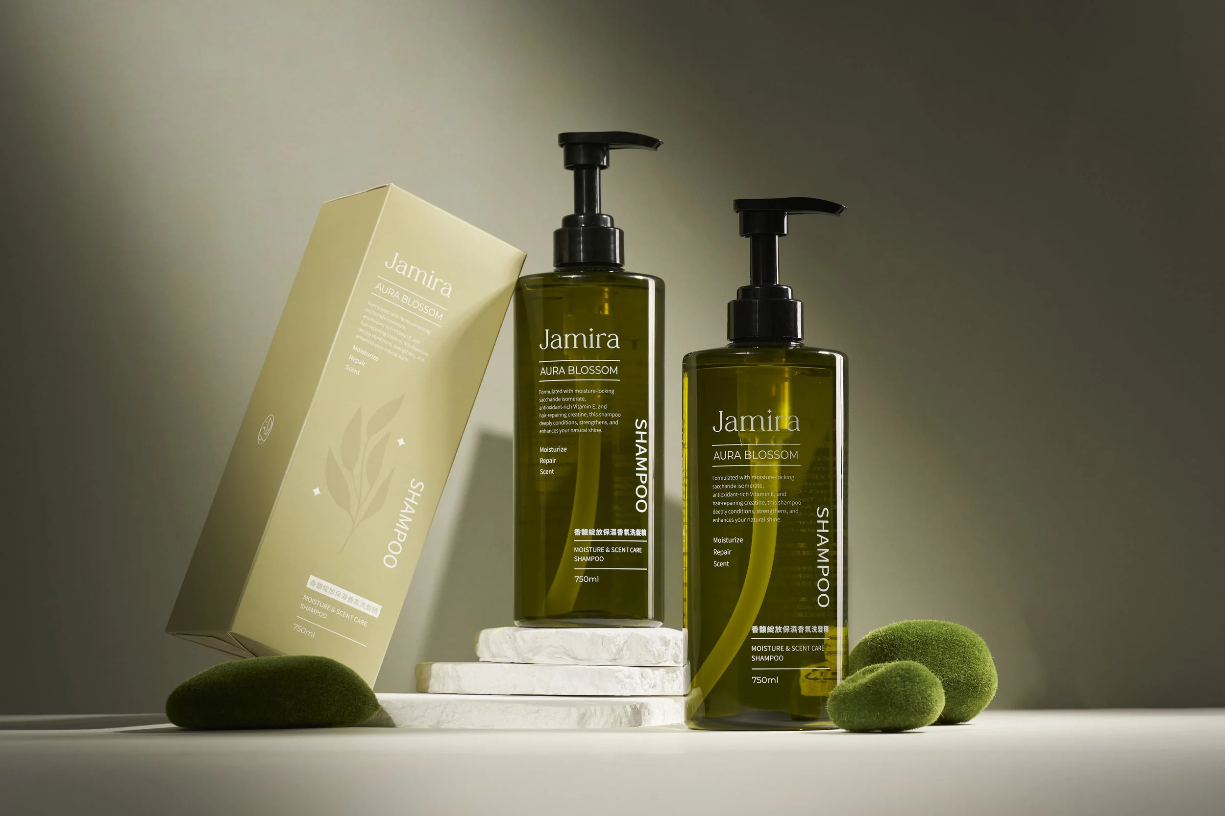 Two bottles of Jamira Aura Blossom shampoo with a beige box, placed on white stone stands, surrounded by small green moss rocks, against a neutral background.