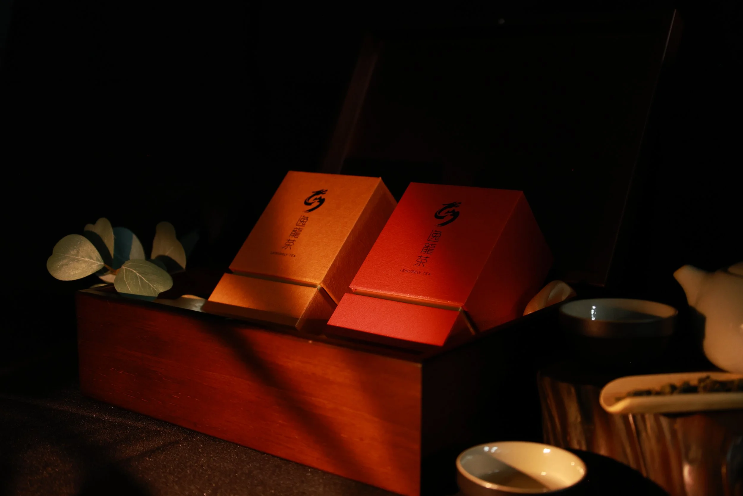 A wooden box contains two orange and red tea boxes with black Chinese characters and symbols, surrounded by tea cups, white flowers, and a dark background.