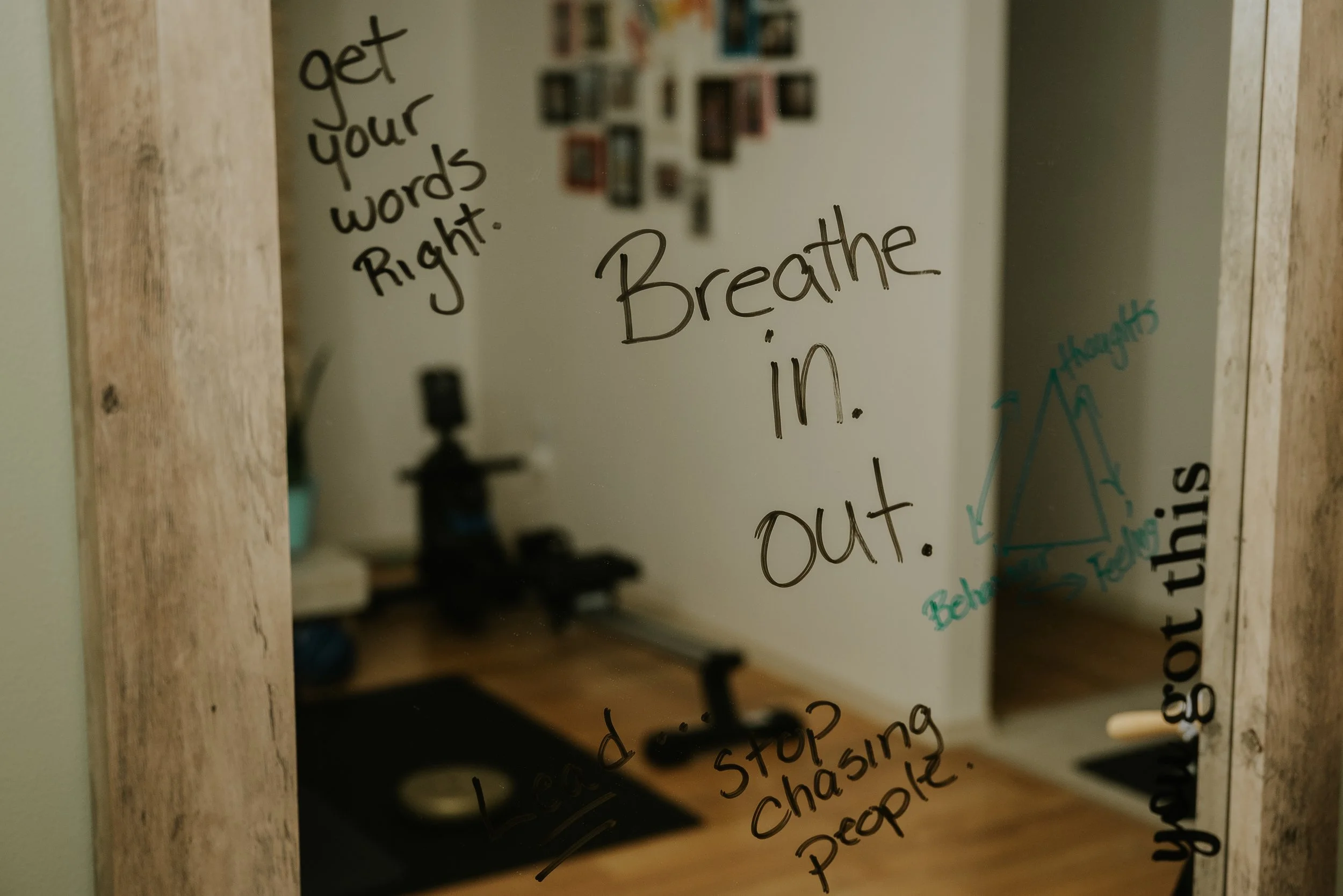 Mirror with motivational and instructional handwritten messages, including 'Breathe in. Out.', 'get your words right.', and 'stop chasing people'. Reflection shows a home gym with a wooden floor, black mats, and some fitness equipment.