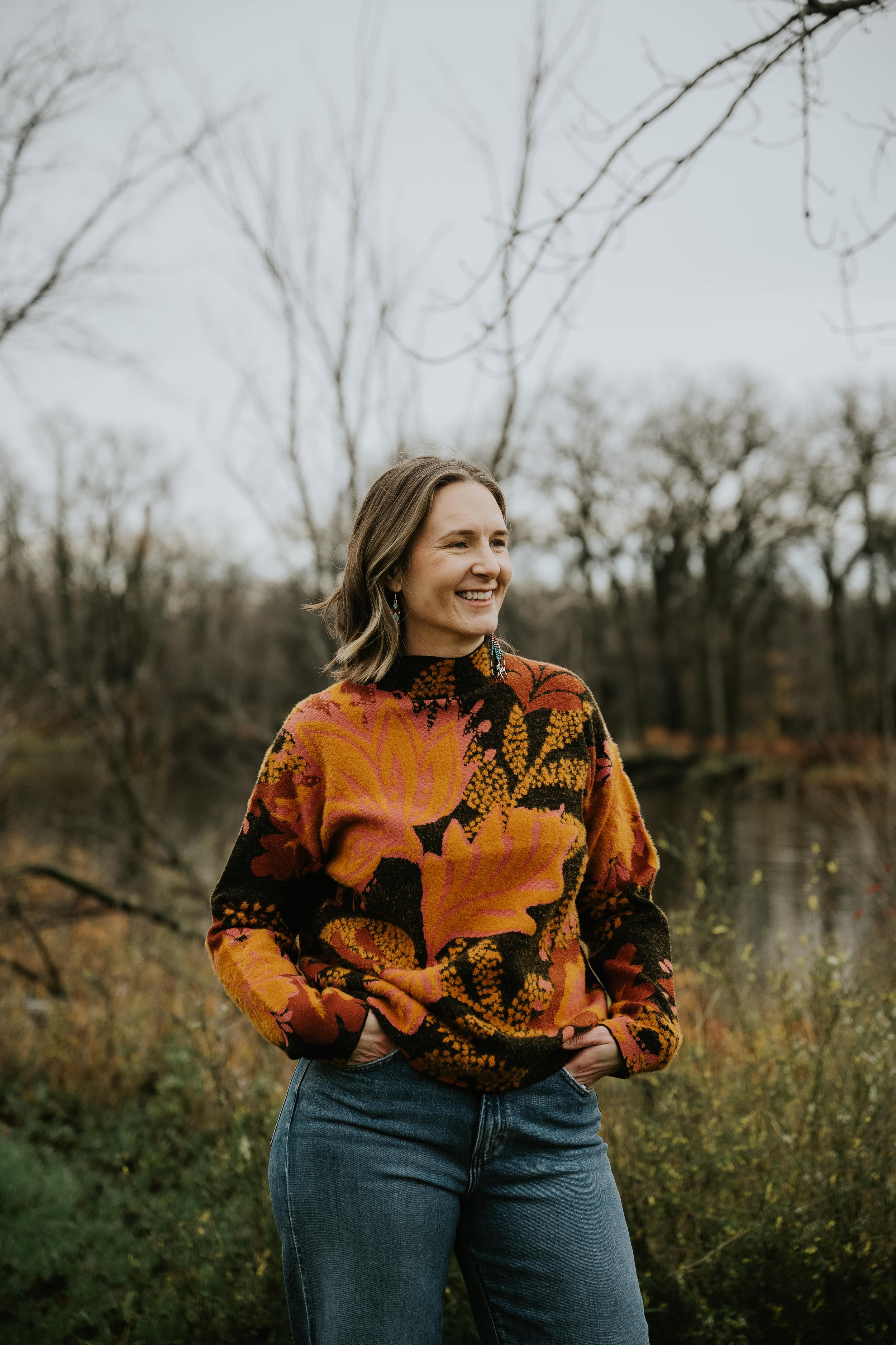 A woman with shoulder-length hair smiling outdoors during fall, wearing a colorful floral-patterned sweater and blue jeans, standing near a lake with leafless trees in the background.