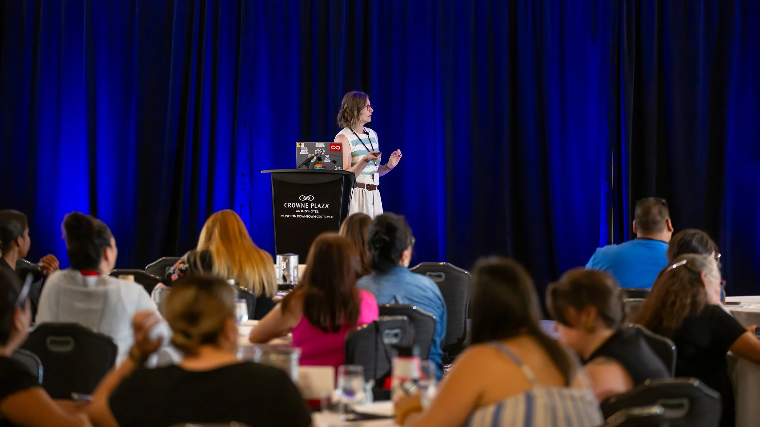 A woman in striped top and white skirt giving a presentation on stage at Crowne Plaza hotel with a laptop, audience members seated at round tables listening.