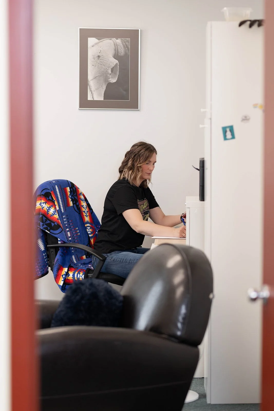 A woman with wavy hair, dressed in a black T-shirt and jeans, is sitting at a desk in an office or waiting room, working on paperwork or a device. Behind her is a framed black-and-white photograph of a whale's head on the white wall. The furniture includes a colorful, patterned jacket draped over the back of her chair and a black leather couch in the foreground.