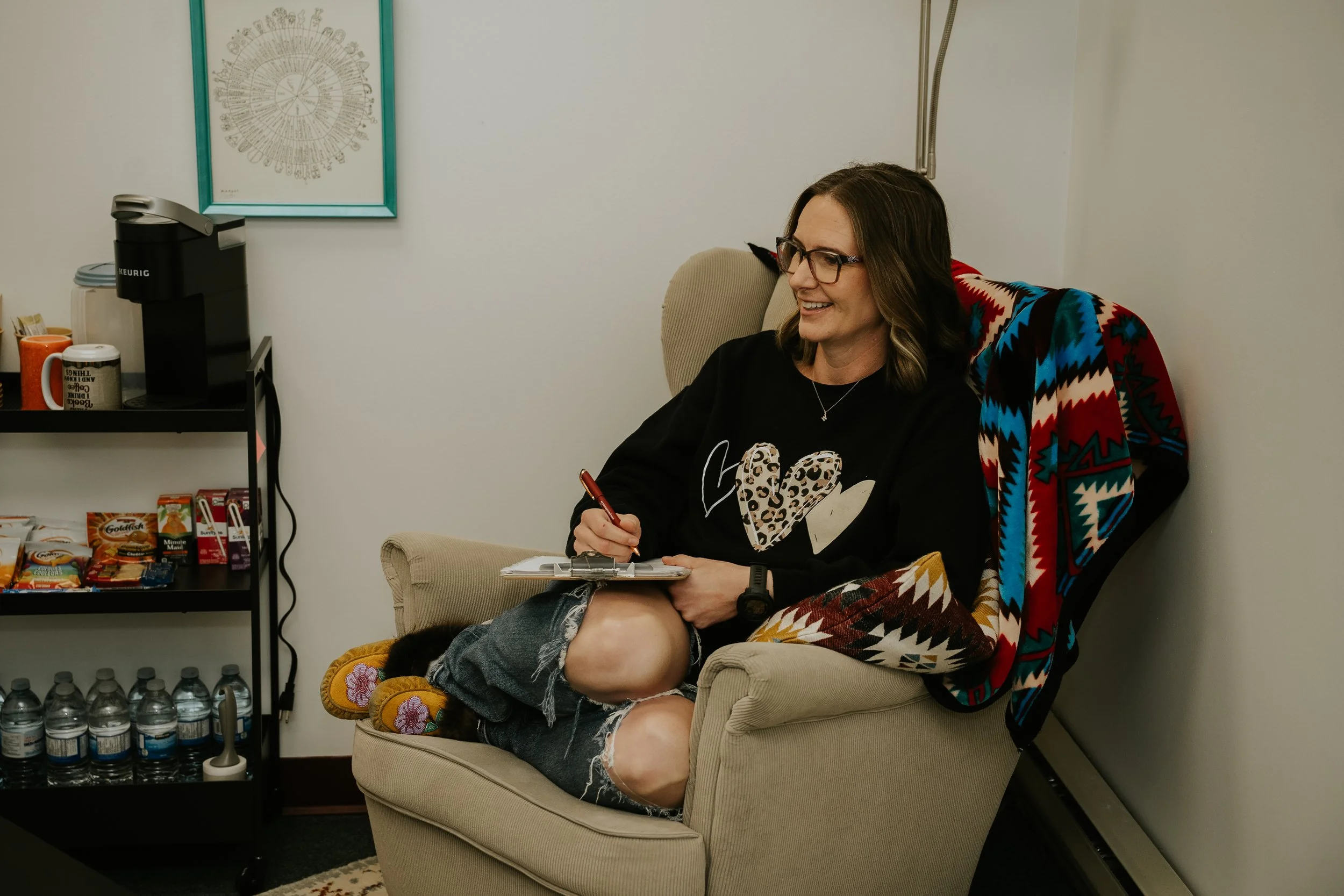 Woman sitting in a beige armchair, smiling, holding a clipboard and pen, wearing glasses, a black shirt with leopard print hearts, and knee-length denim shorts, in a room with a snack cart and water bottles.