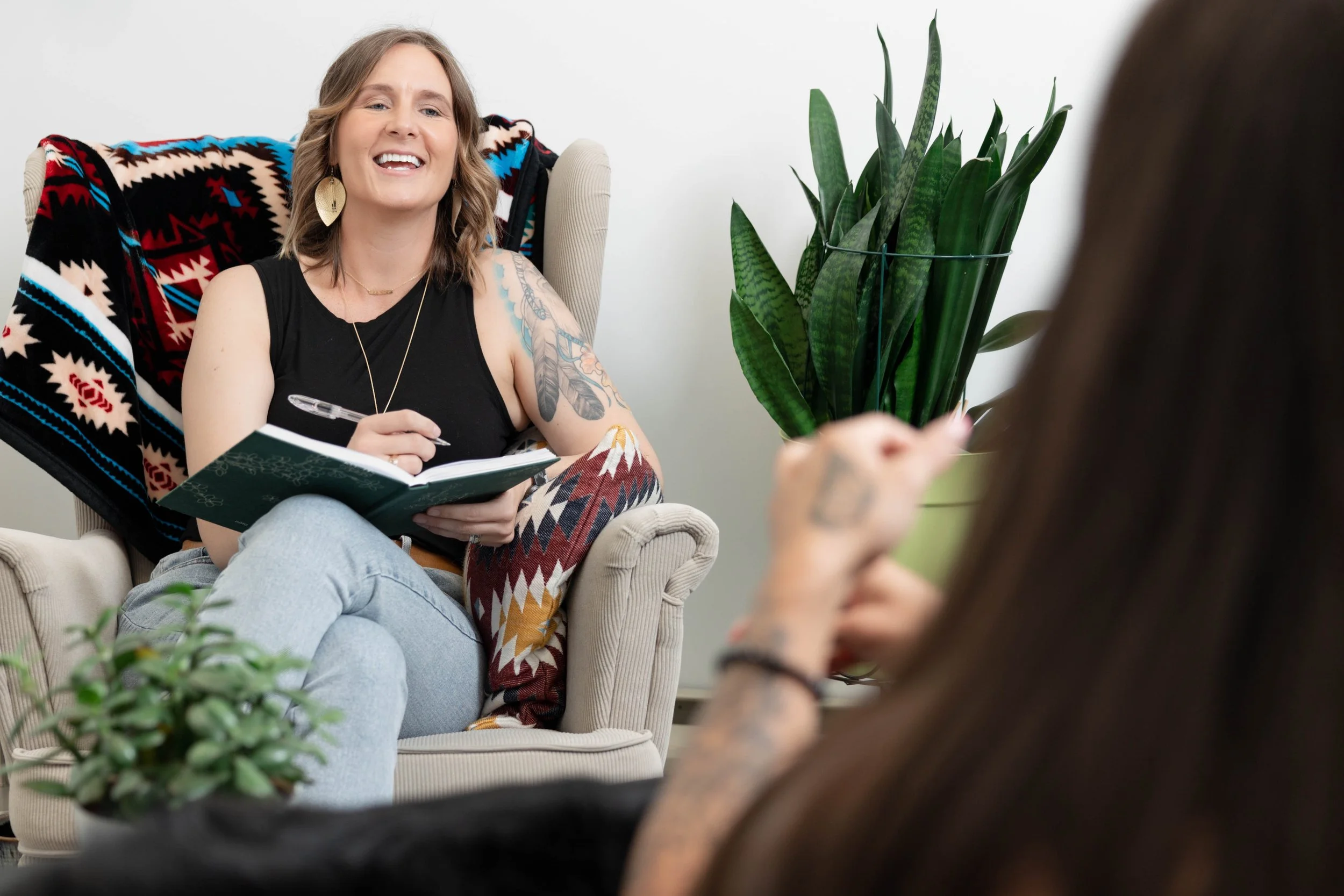 Woman with shoulder-length hair, tattoos, and earrings smiling, sitting on a beige armchair with a notebook, during a conversation with another person, with a large green plant beside her.