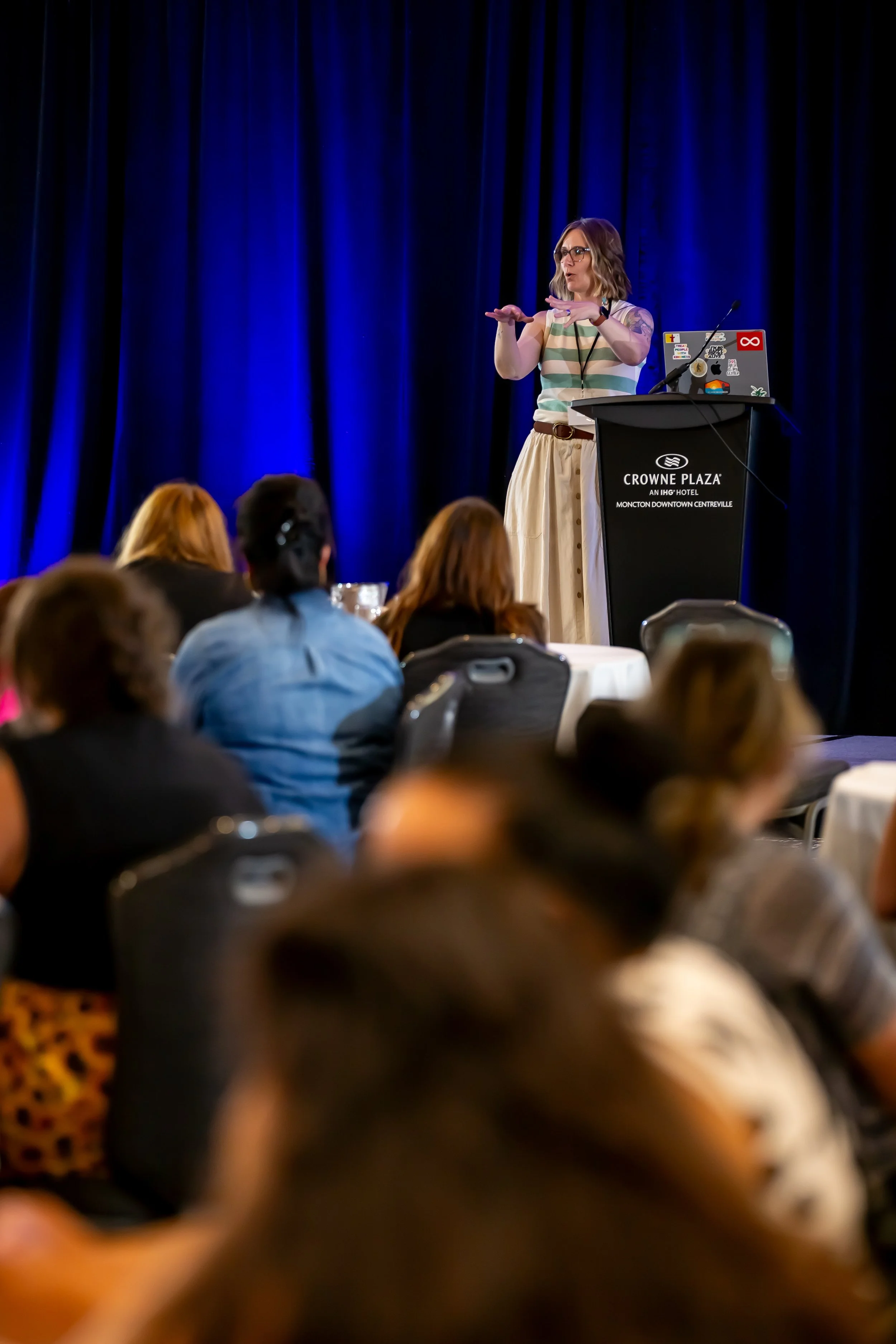 A woman giving a presentation on stage at Crowne Plaza hotel to an audience seated at round tables.