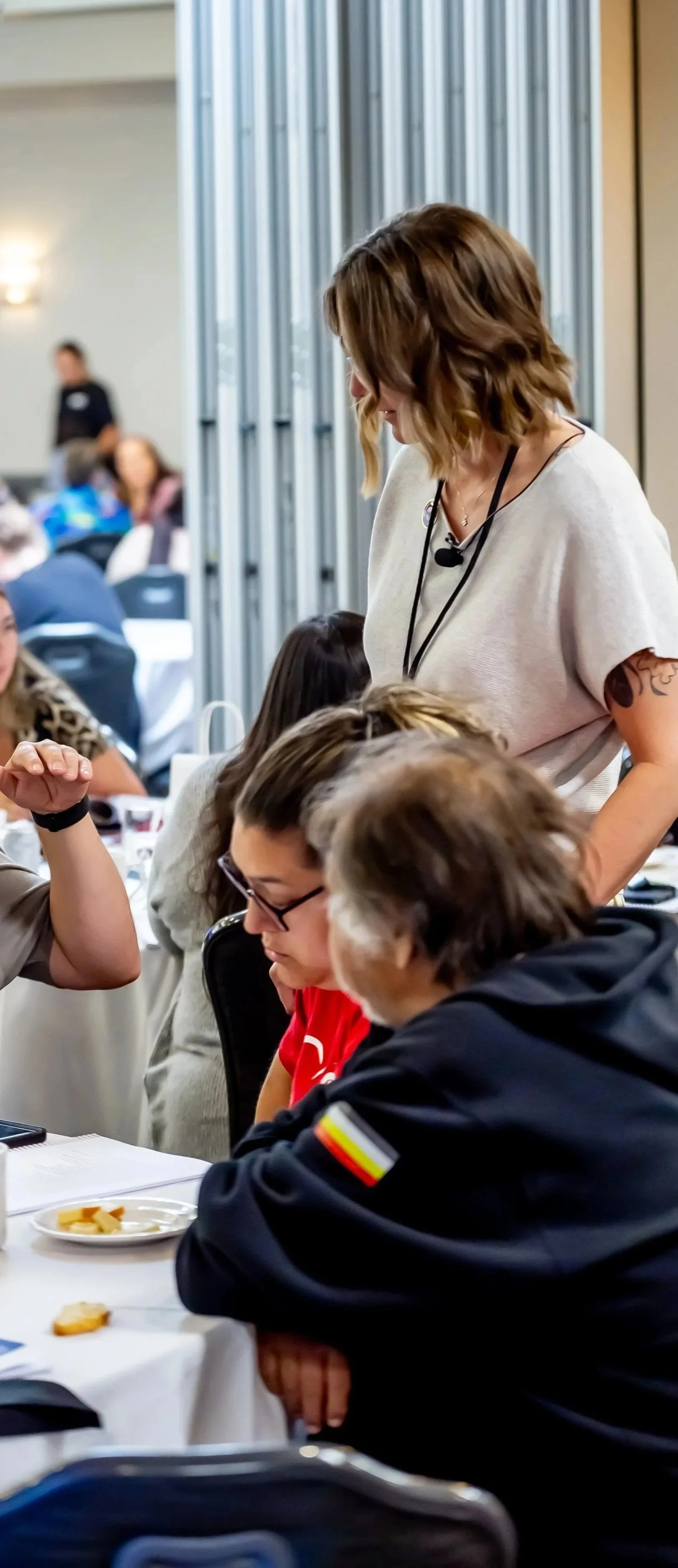 A woman with shoulder-length brown hair and tattoos on her arm is standing and talking to two seated women at a round table during a conference or workshop in a large, well-lit room.