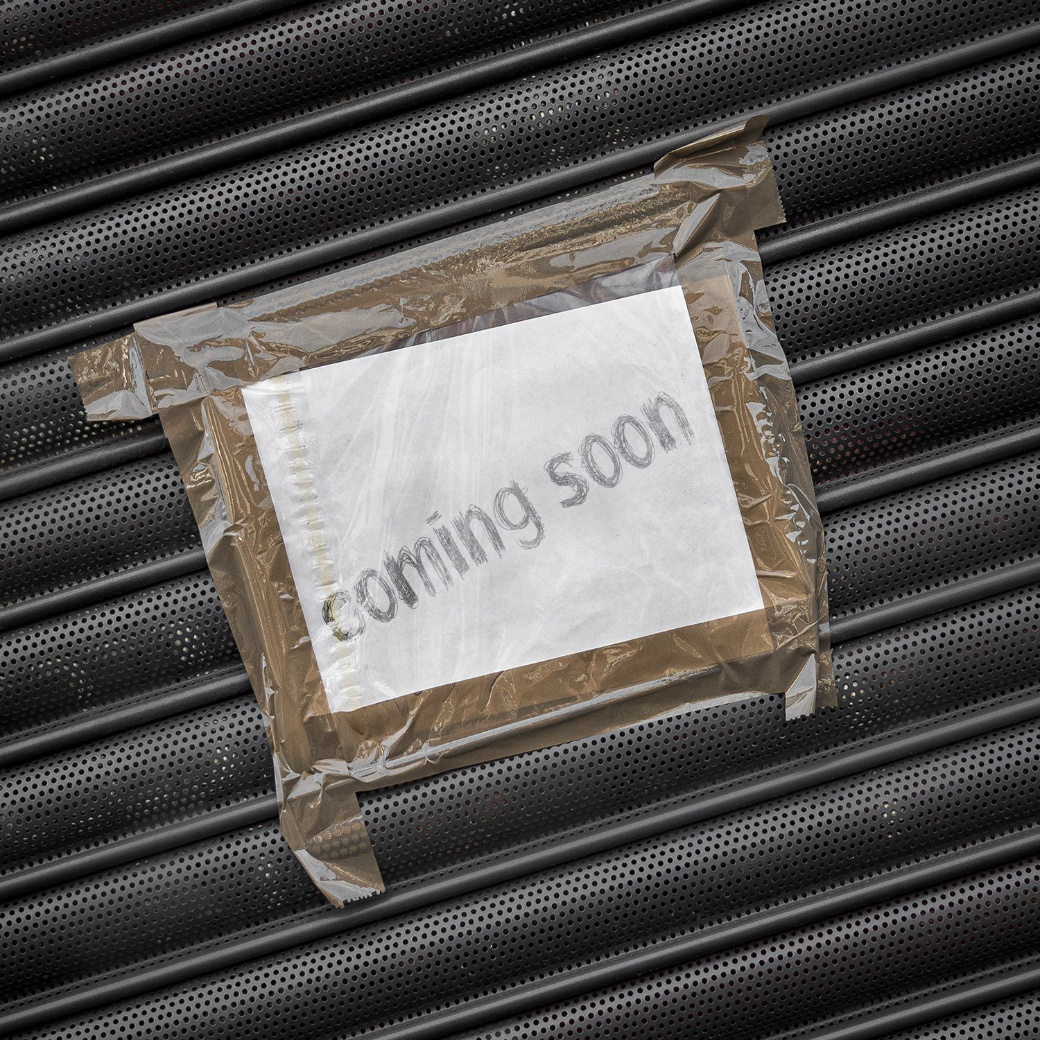 A white piece of paper with the words "coming soon" taped to a perforated metallic surface, covered with transparent plastic.