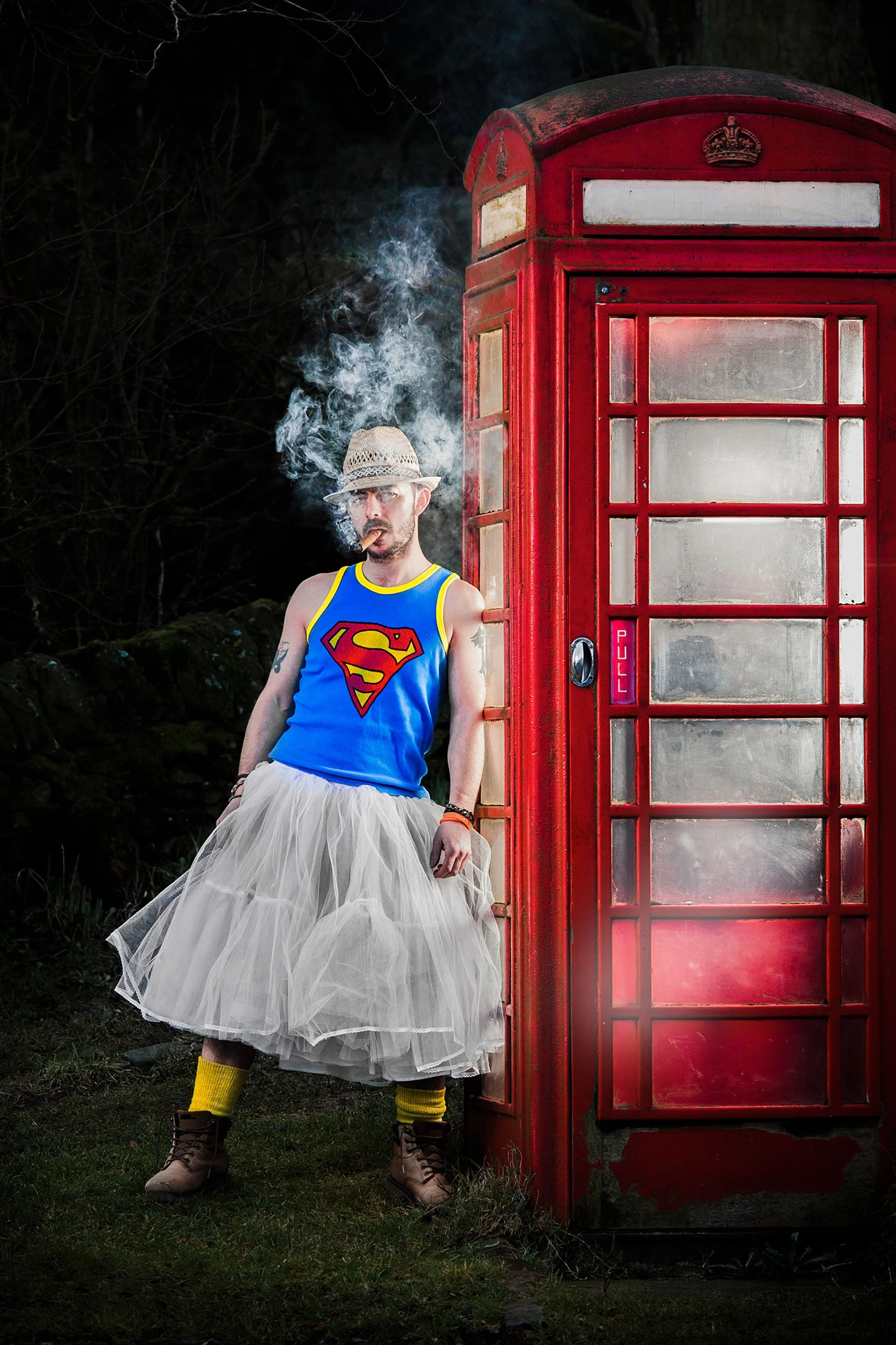 Fashion photograph of male model wearing a Superman tank top, tutu, and hat, smoking, leaning against a red British phone booth. Photography by Dave Wall Photo