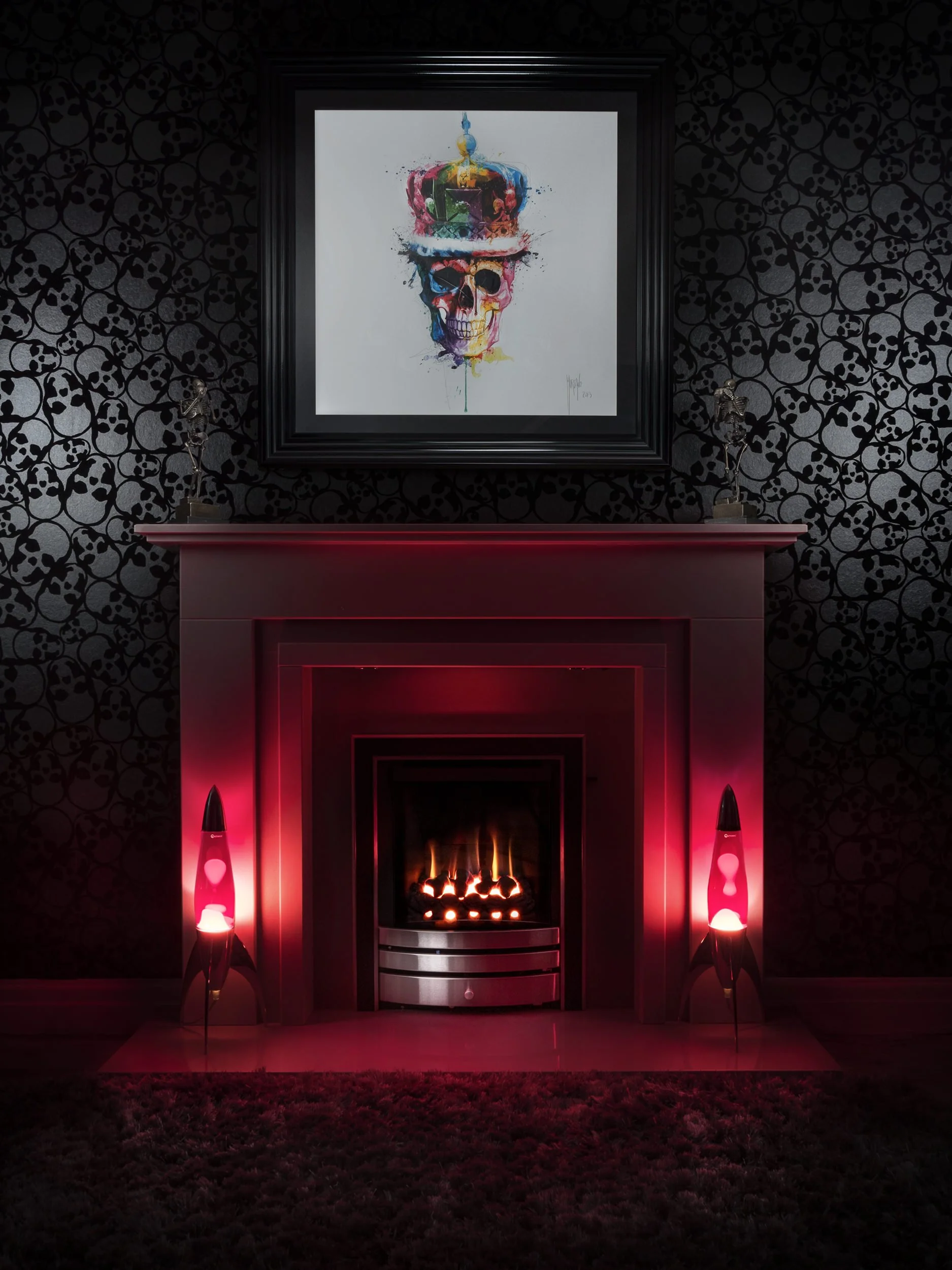 House interior with a modern fireplace featuring two red lava lamps. Above the mantel is a colorful skull painting wearing a crown. The wallpaper has a subtle pattern of skulls. Photography by Dave Wall Photo