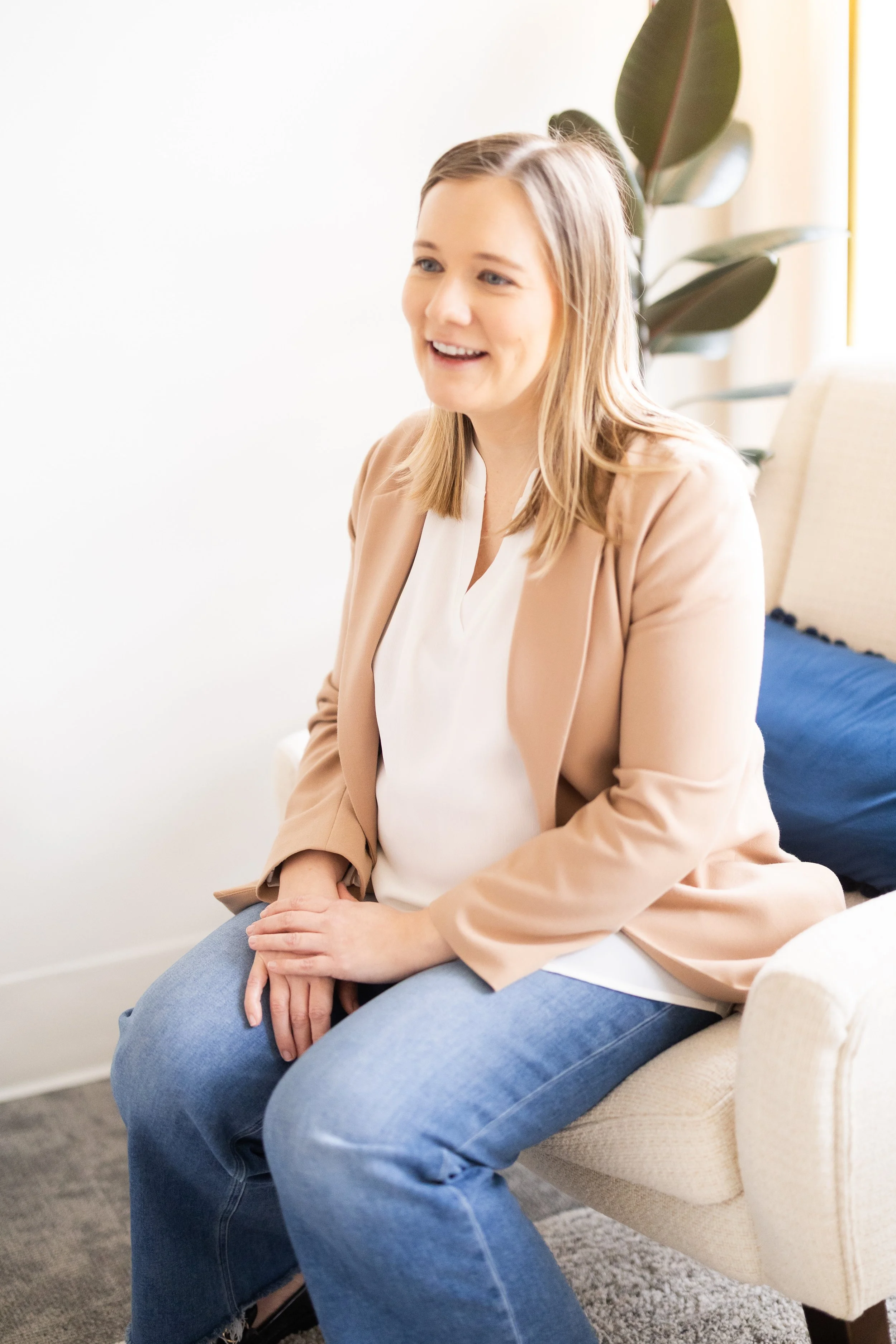 Therapist for teens in Bellevue supporting confidence, boundaries, and emotional health, seated on a beige chair, smiling, wearing a beige blazer, white blouse, and blue jeans, with a plant in the background.
