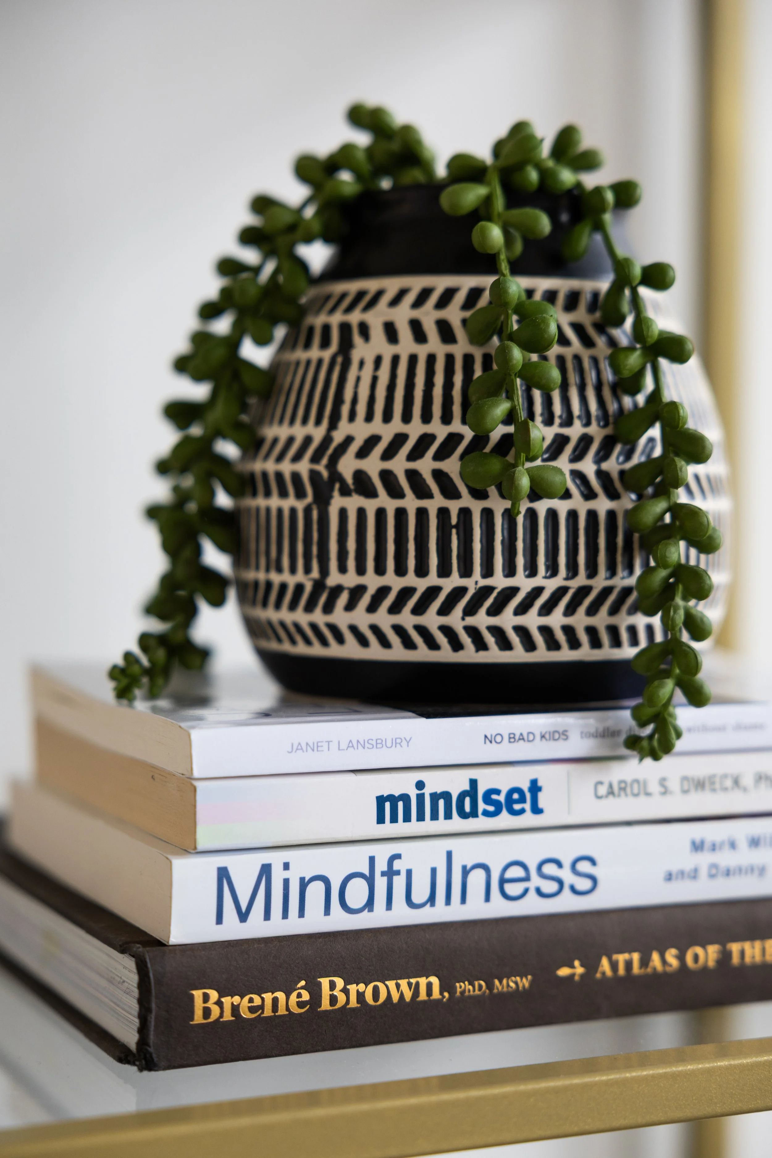 A black and white patterned vase with trailing green succulent plant, placed on a stack of four books on a gold table, with titles about mindfulness and psychology, Alignwell Modern Therapy's in-person counseling office in Bellevue, Washington.