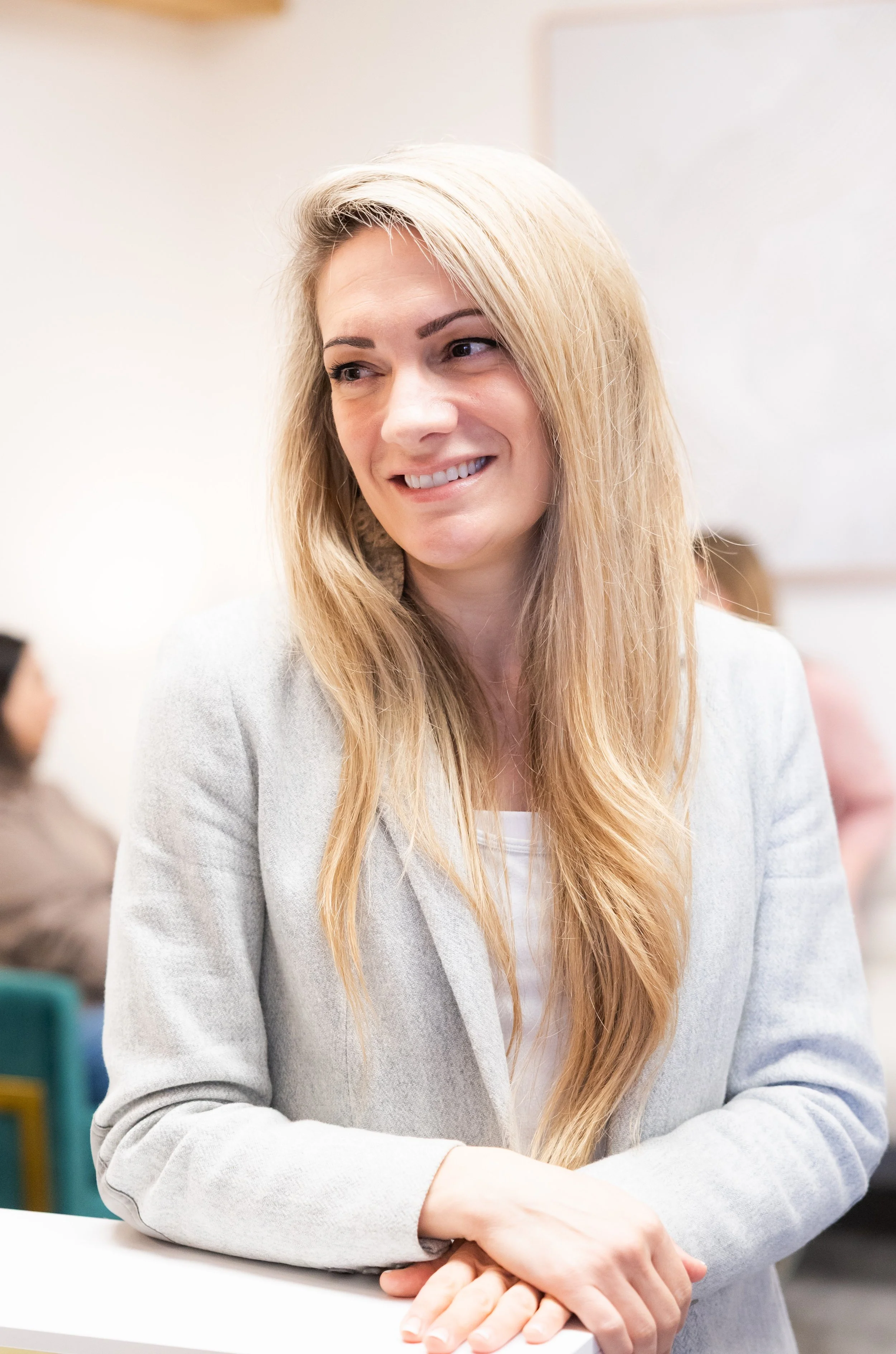 Therapist in Bellevue specializing in neurodivergent adults, wearing a grey blazer, smiling and leaning on a white surface, in an indoor setting with other people in the background.