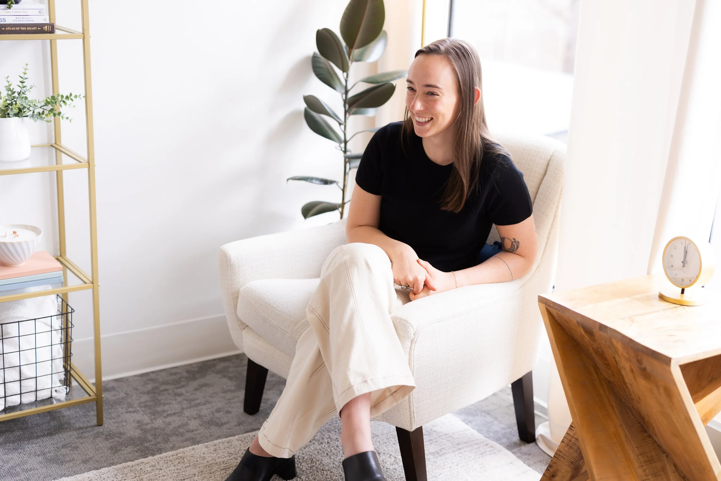 Bellevue couples therapist supporting premarital counseling and long-term partnership growth, sitting in a light-colored armchair, smiling, wearing a black t-shirt, cream-colored pants, and black shoes
