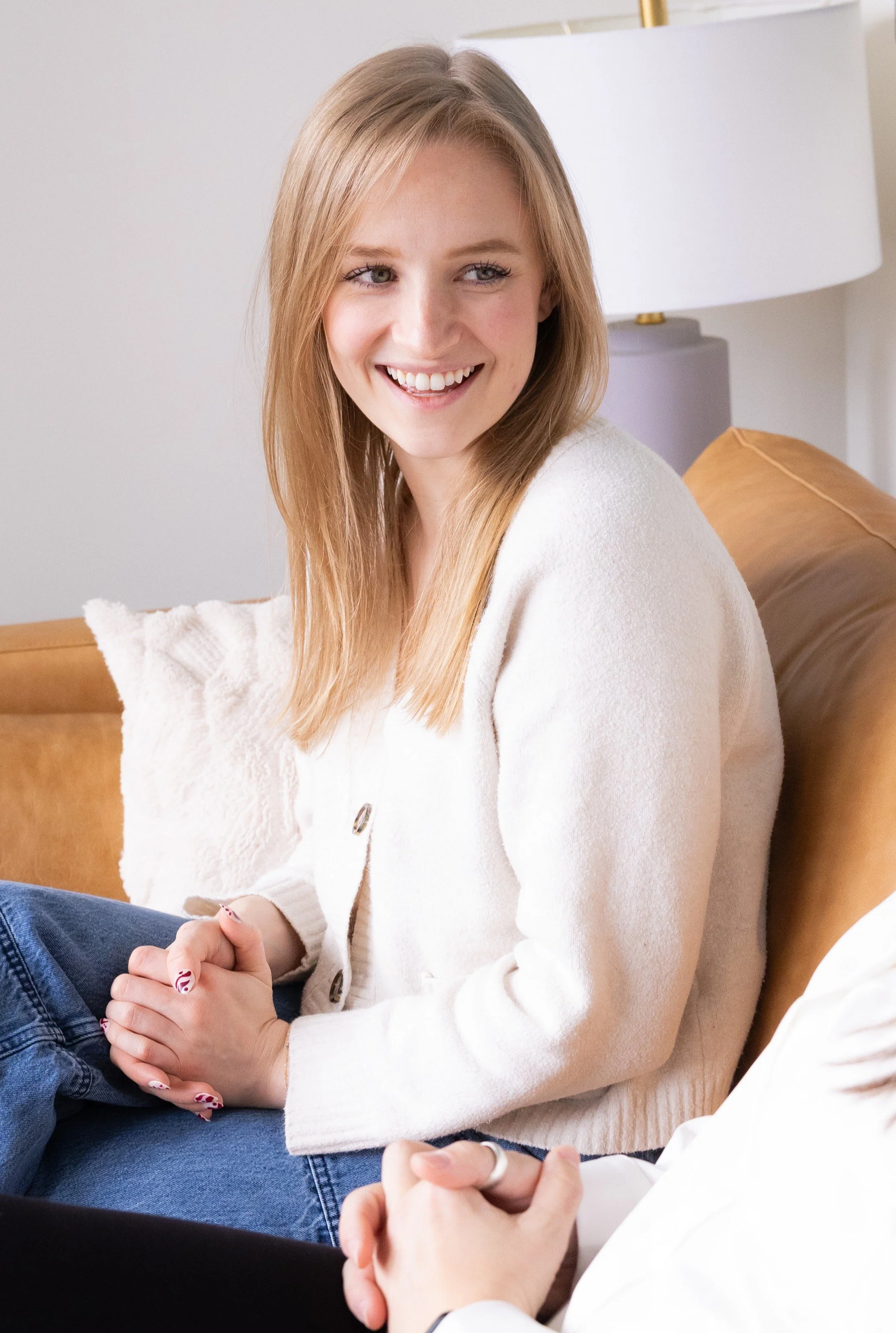 Modern therapist for anxiety in Bellevue, Washington, smiling, sitting on a couch indoors, wearing a white cardigan and blue jeans, with her hands clasped, engaged in conversation.