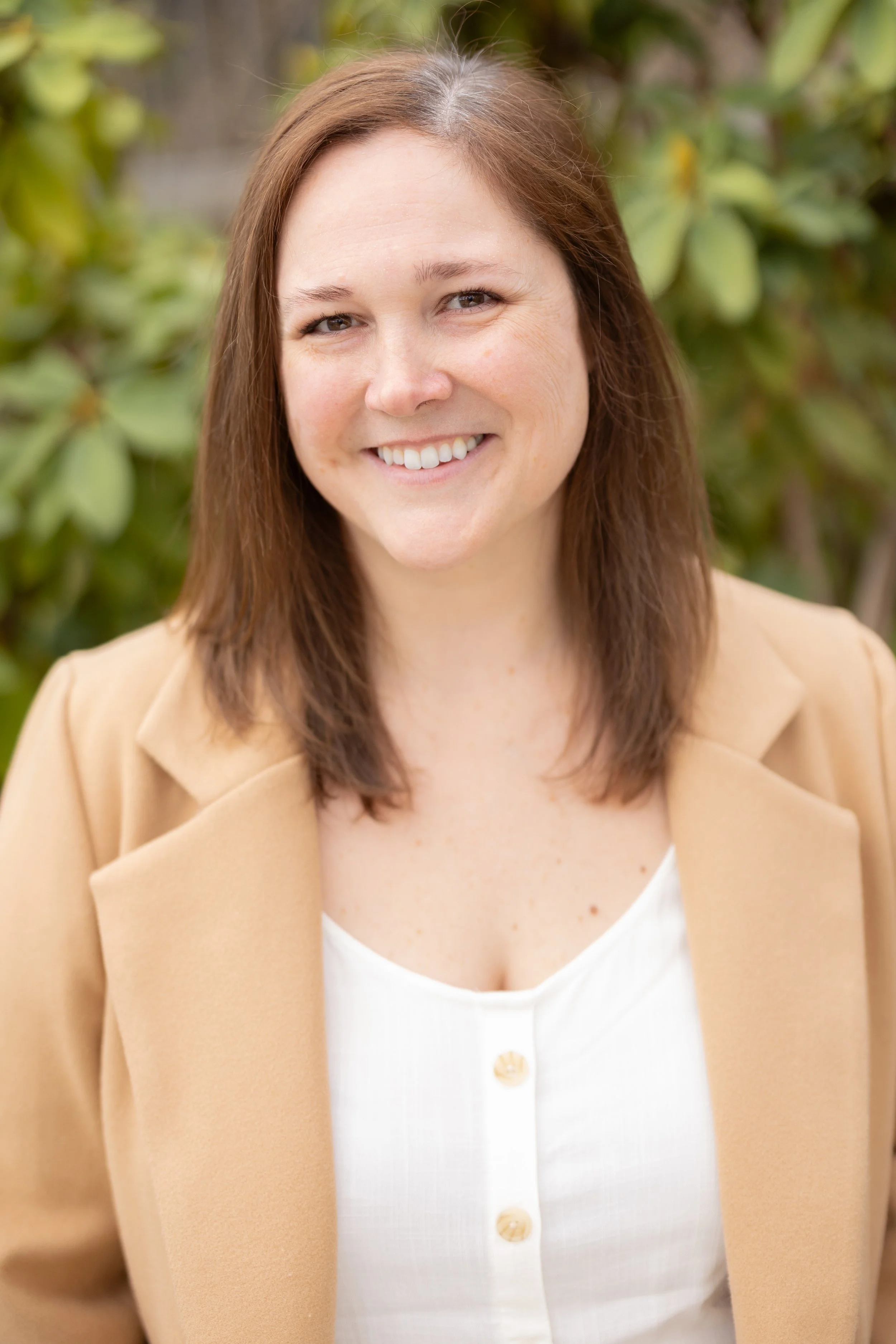 A smiling woman with shoulder-length brown hair, wearing a beige blazer over a white top, standing outdoors with green foliage in the background.