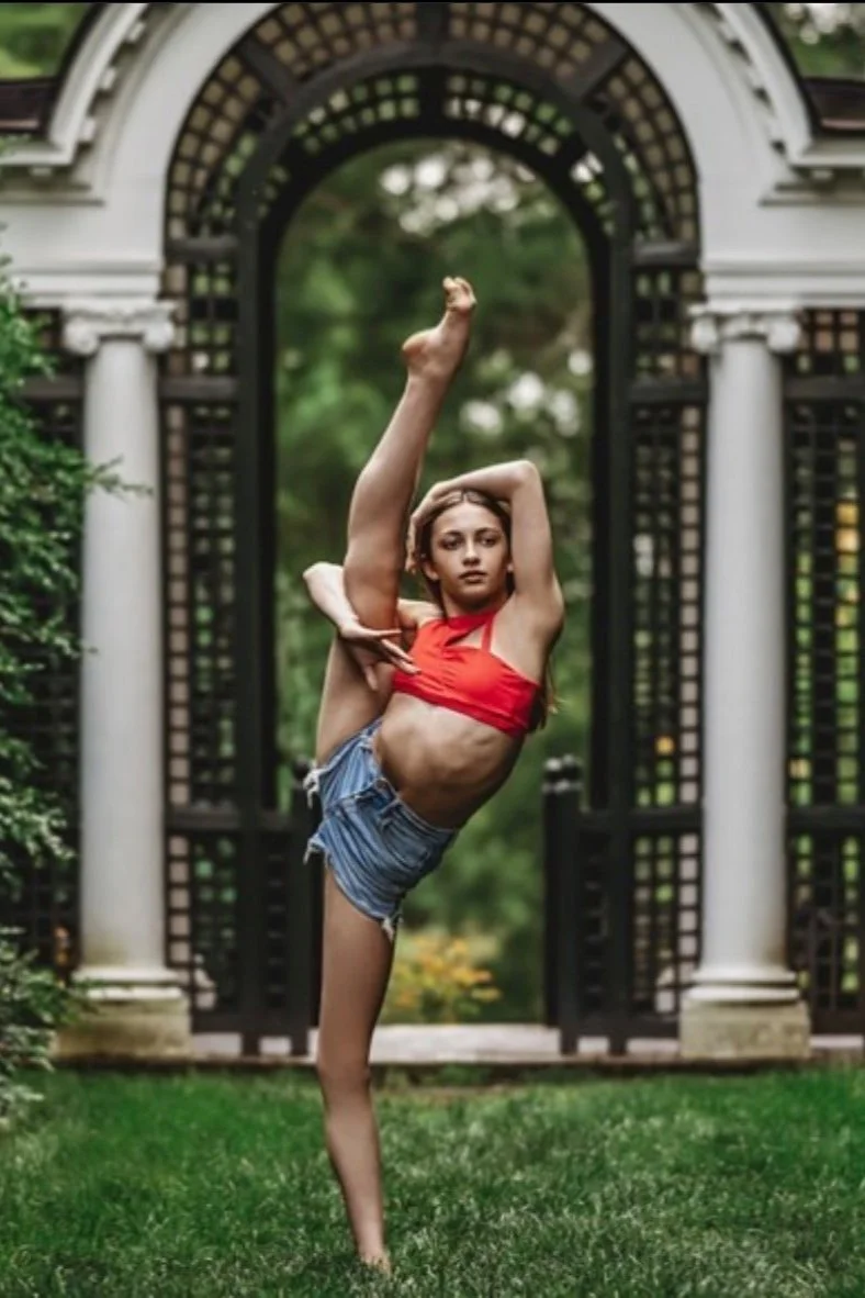 Person performing a standing split in a garden, wearing a red top and denim shorts, with an ornate archway in the background.