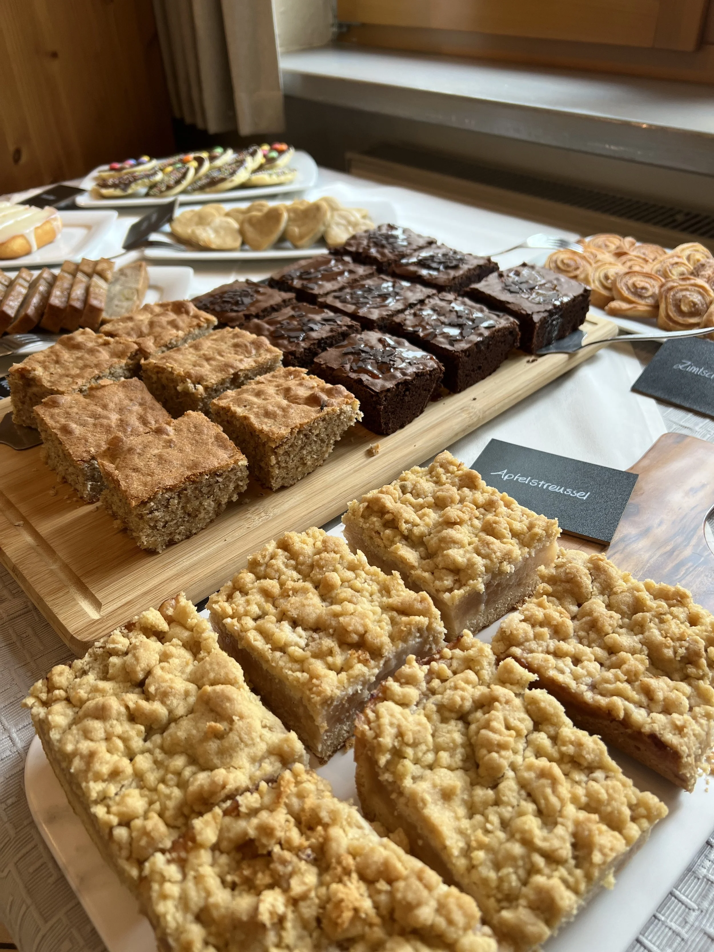 Verschiedene Kuchen und Gebäck auf Tellern, darunter Streuselkuchen, Schokokuchen, Plätzchen und Croissants, auf einem Tisch im Raum.