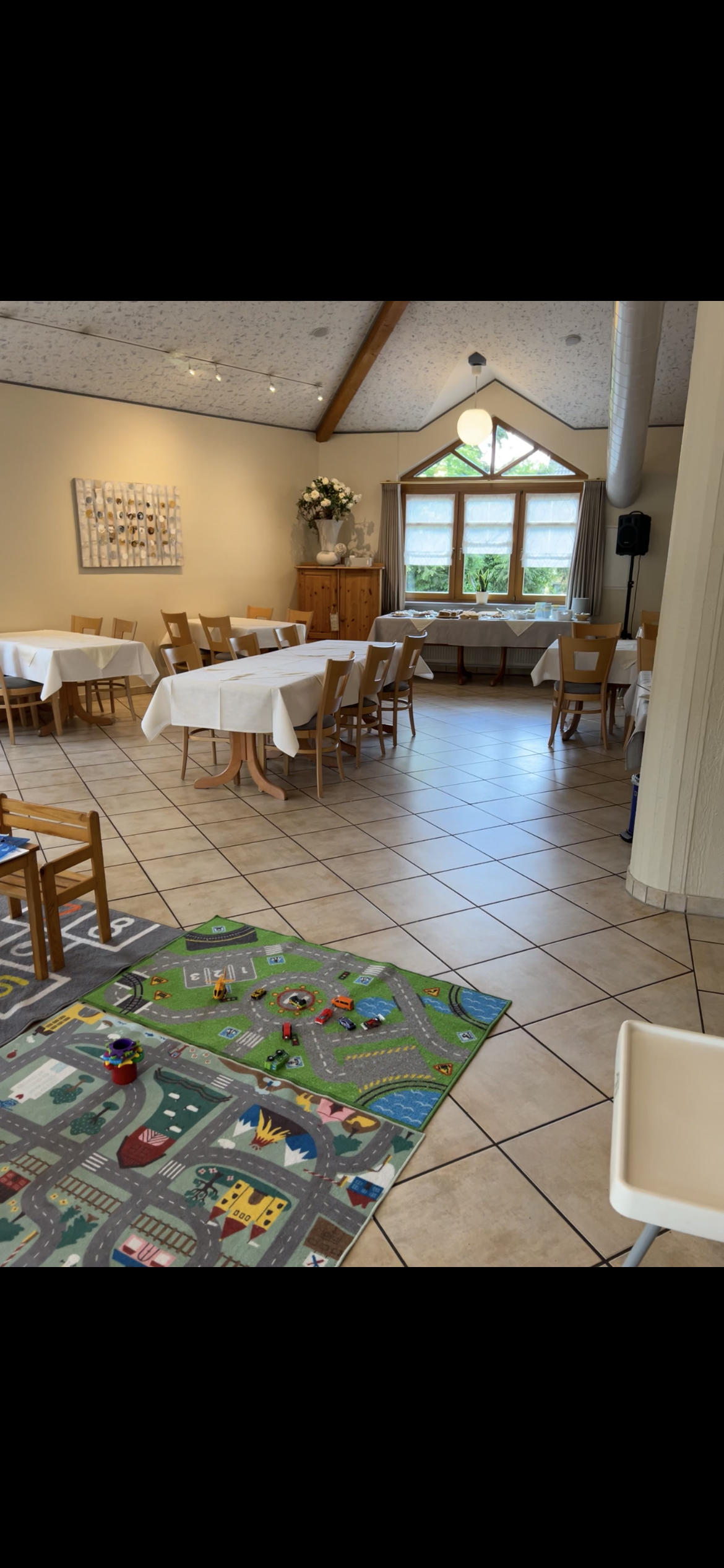 Ein Esszimmer mit mehreren gedeckten Tischen, einem Klavier, einem Blumenstrauß und einem Fenster mit Vorhängen. Im Vordergrund befindet sich ein Kinderspielteppich mit Spielzeug.