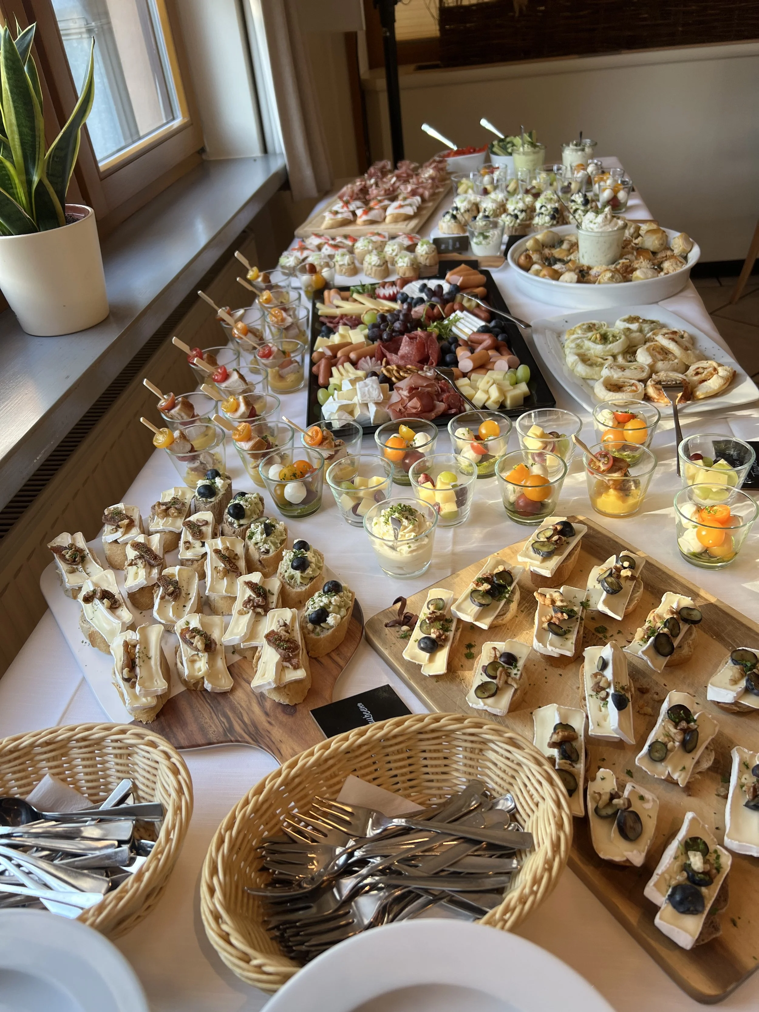 Büffet mit verschiedenen Häppchen, Käse, Wurst, Obst und Desserts. Es gibt auch zwei Körbe mit Besteck und Servietten.
