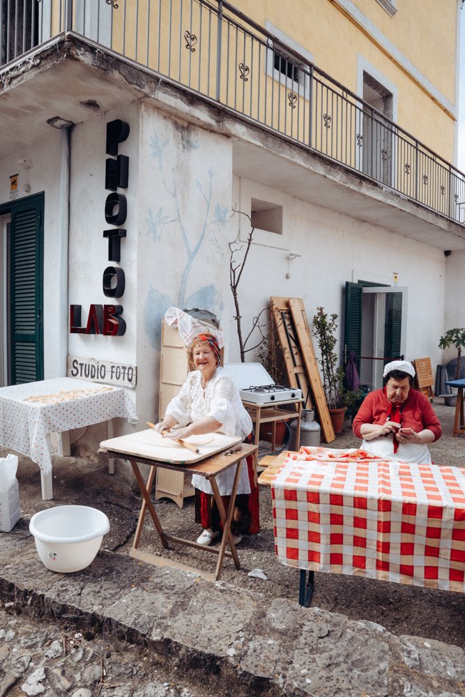 Due donne che cucinano all'aperto, una che sorride e una che guarda il cellulare, con un edificio sullo sfondo e un tappeto a scacchi rosso e bianco sulla tavola.