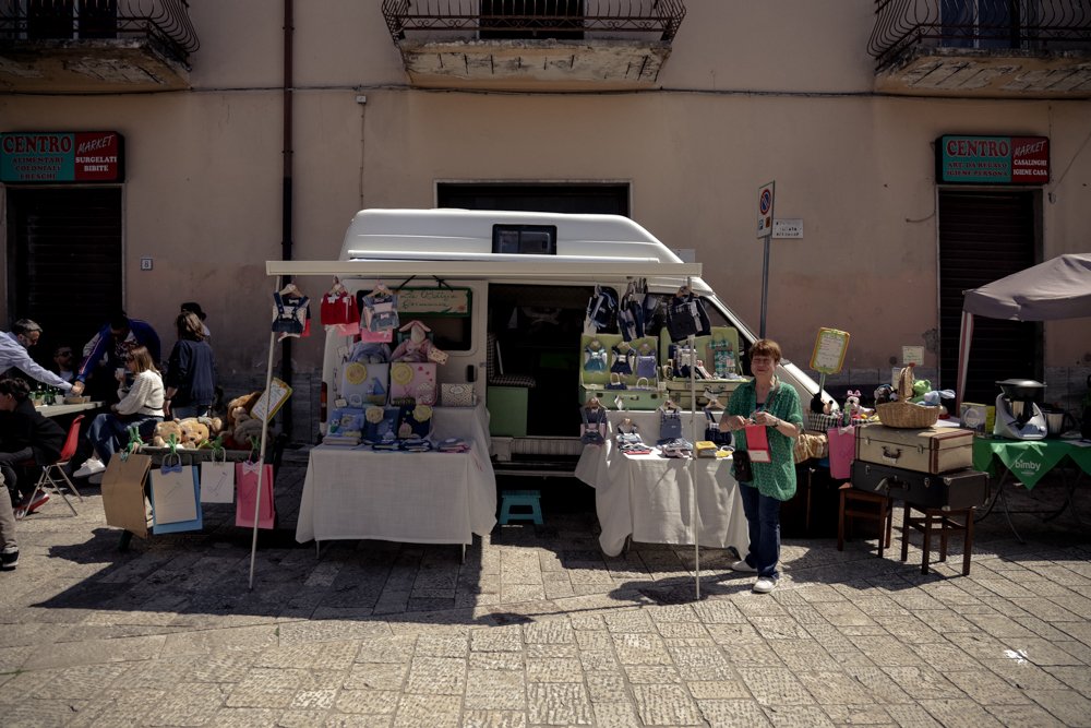 Bancarella di strada con venditore, oggetti artigianali, peluche e accessori esposti davanti a un furgone bianco. Persone sedute a un tavolo sulla sinistra.