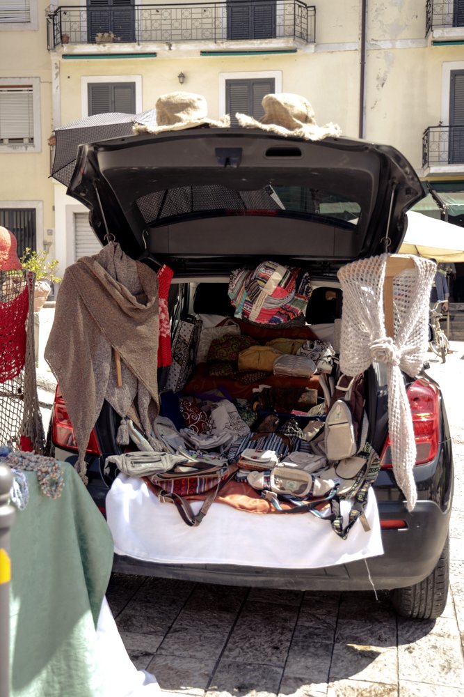 Macchina usata come bancarella, carica di borse, cappelli e tessuti, in strada di città.