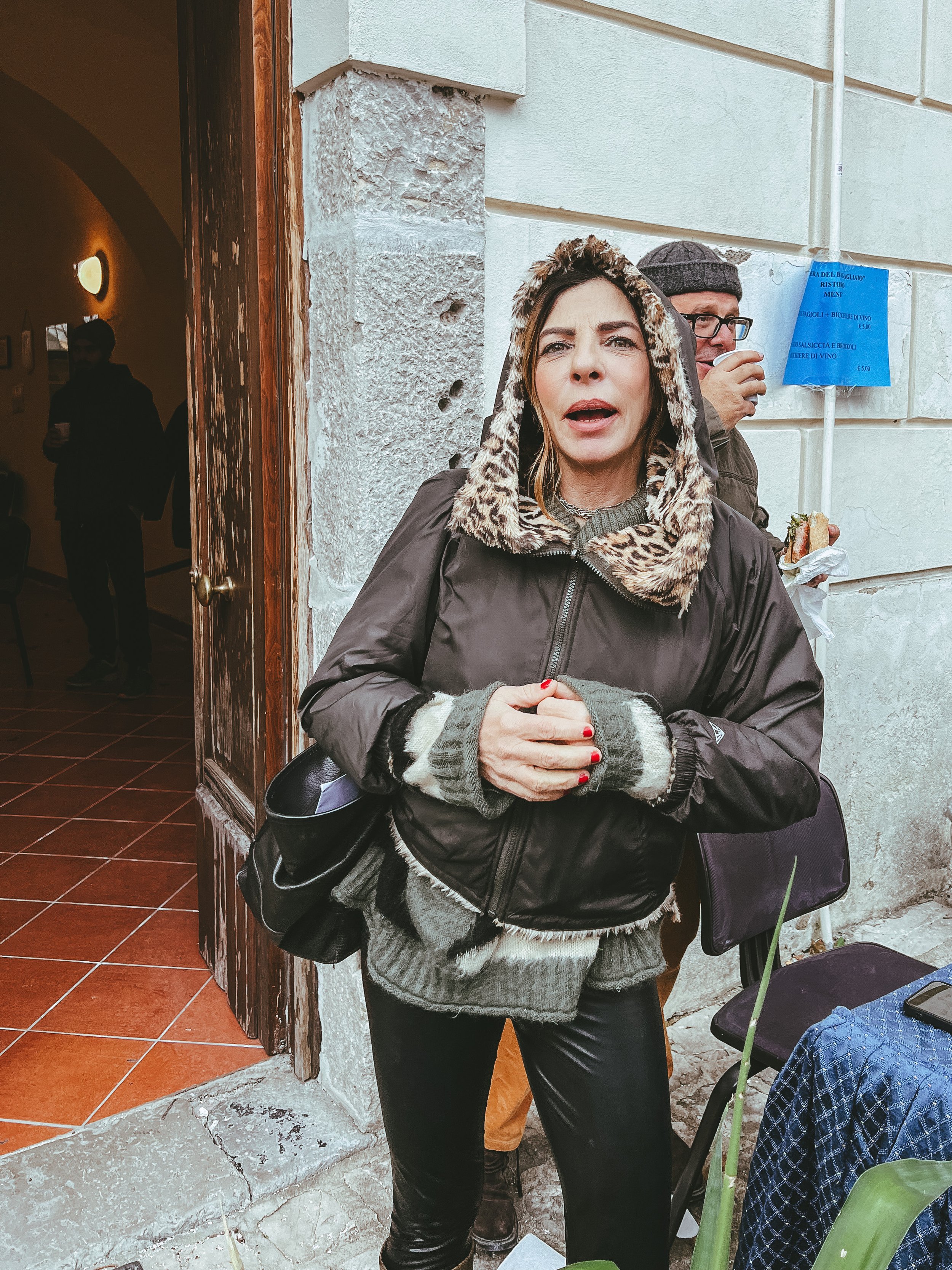 Due persone fuori da un edificio; una donna con giacca e cappuccio leopardato, un uomo dietro di lei con cappello, entrambi vicino a una porta aperta.