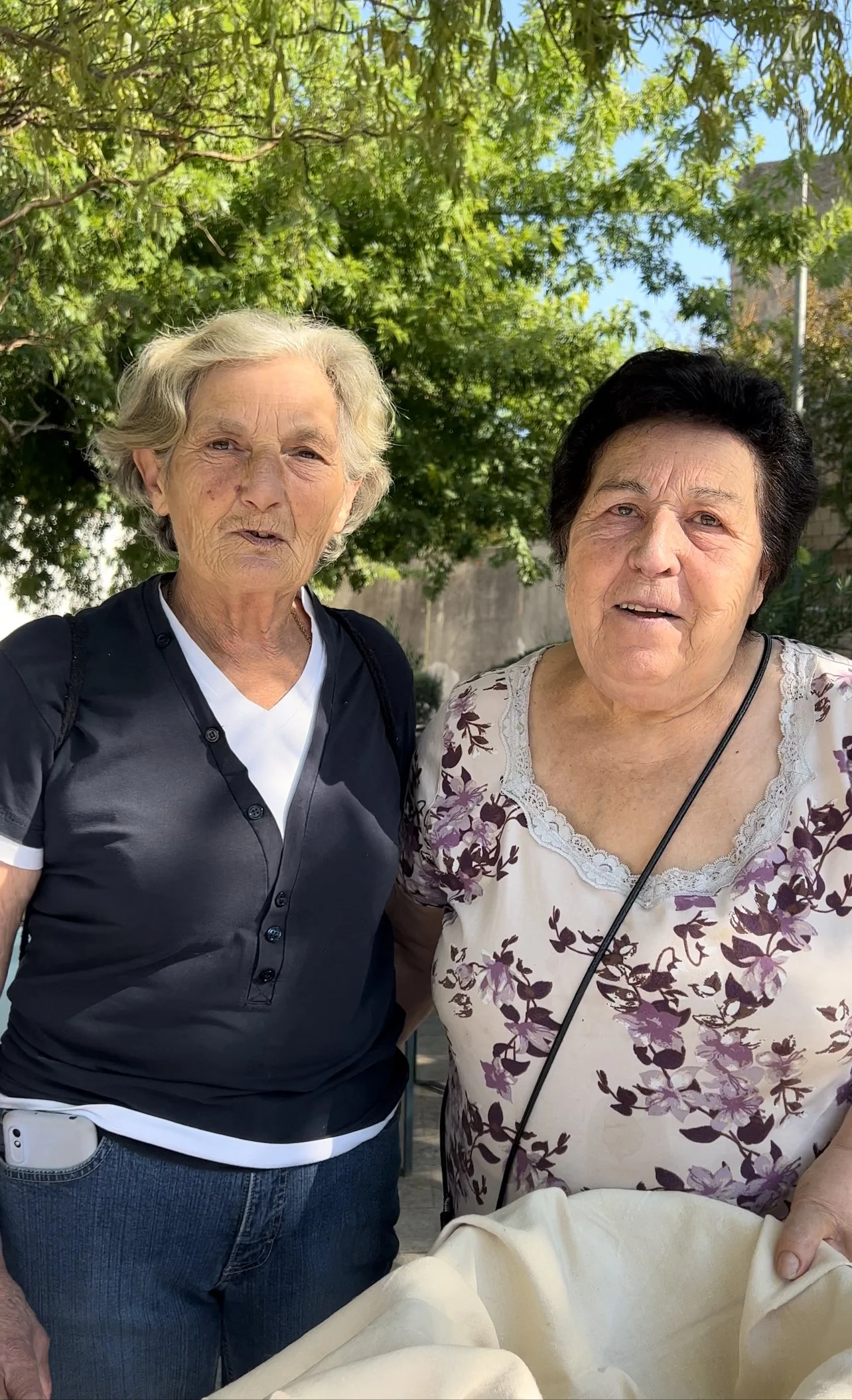 Due donne anziane sorridenti all'aperto, circondate da alberi verdi.