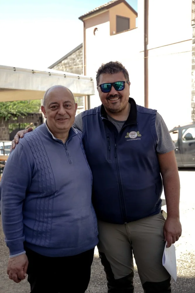 Due uomini sorridenti in piedi all'esterno, uno con occhiali da sole e giacca blu, l'altro con maglione blu. Entrambi sembrano felici e uno tiene un foglio di carta.