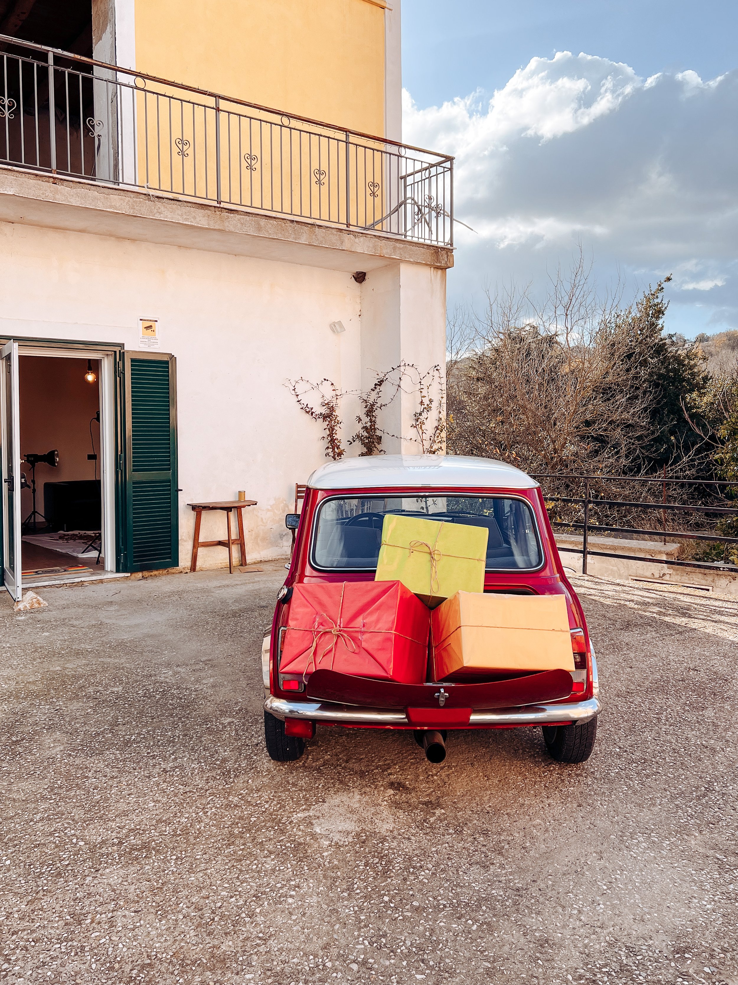 Auto d'epoca rossa con bagagliaio aperto e tre grandi pacchi colorati sul retro, parcheggiata di fronte a una casa con balcone in un'area rurale.