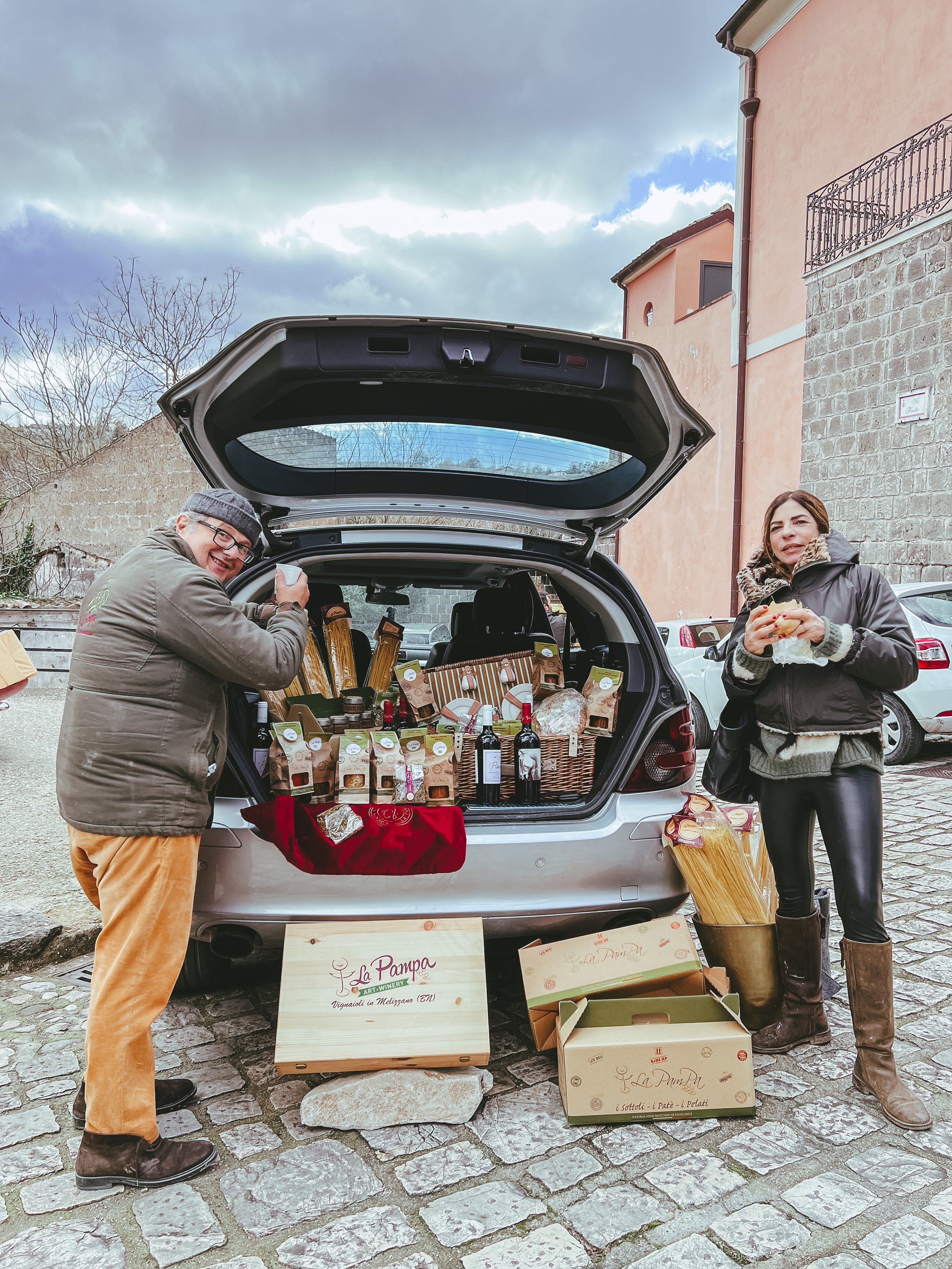 Due persone stanno accanto al bagagliaio aperto di un'auto, che è pieno di prodotti alimentari e bottiglie di vino. Sul pavimento ci sono scatole con scritto 'La Pampa'. Sullo sfondo si vede una strada lastricata e alcuni edifici.