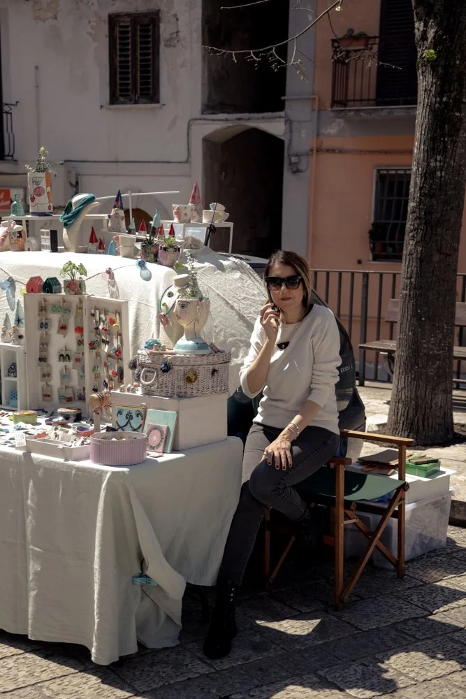 Donna seduta vicino a un banco di artigianato all'aperto, con oggetti colorati esposti su un tavolo coperto da un telo bianco.