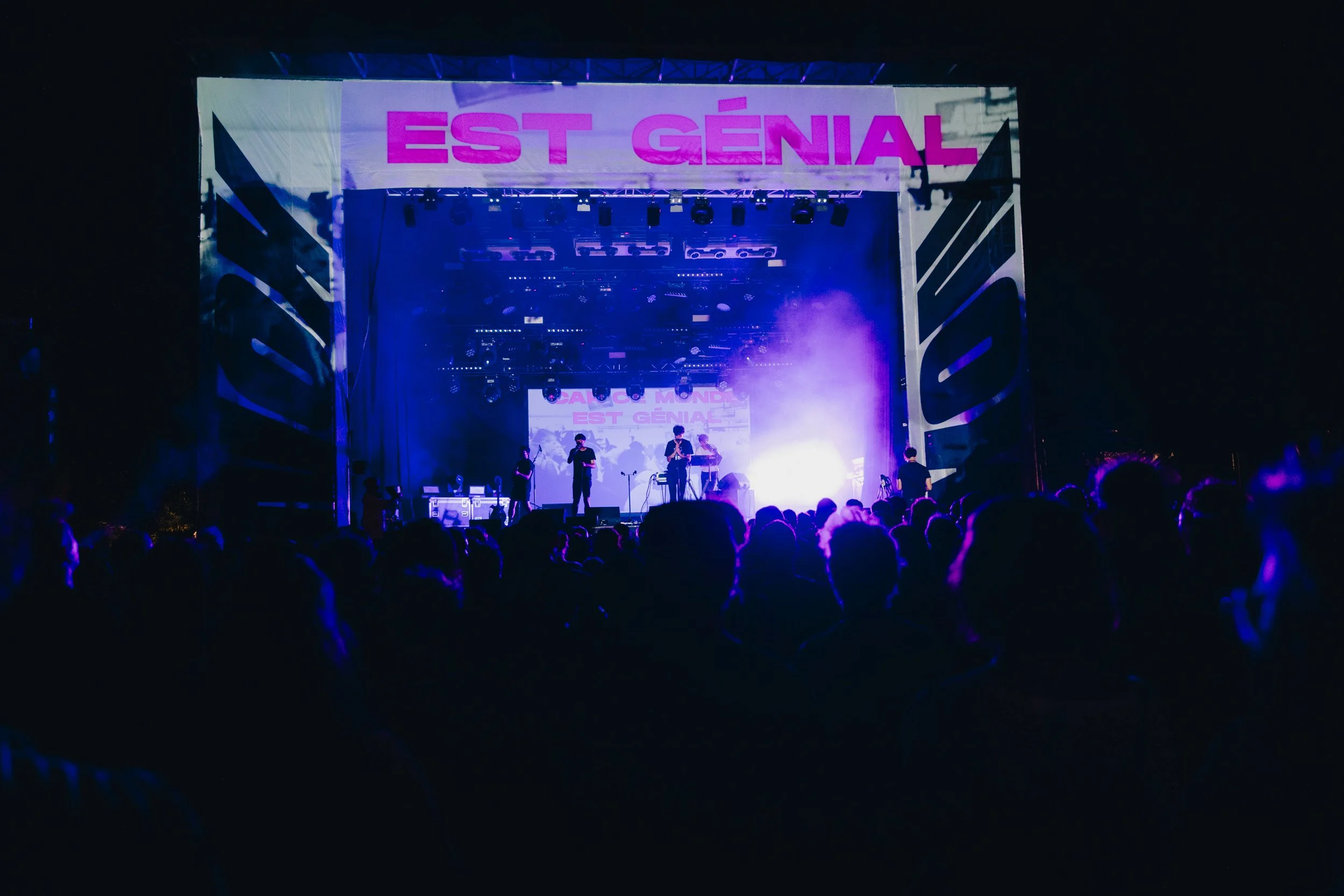 Concert en plein air avec une scène illuminée en bleu et violet, devant un public. Une banderole avec écrit "EST GÉNIAL" est visible en haut de la scène.