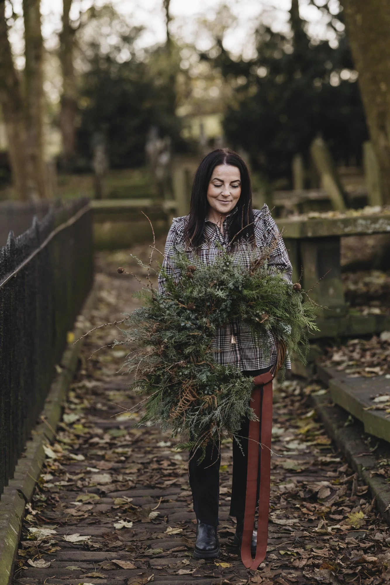 A woman with black hair wearing a checked shirt and black boots walking on a leaf-covered pathway in a park, holding a large bunch of green forest greenery.