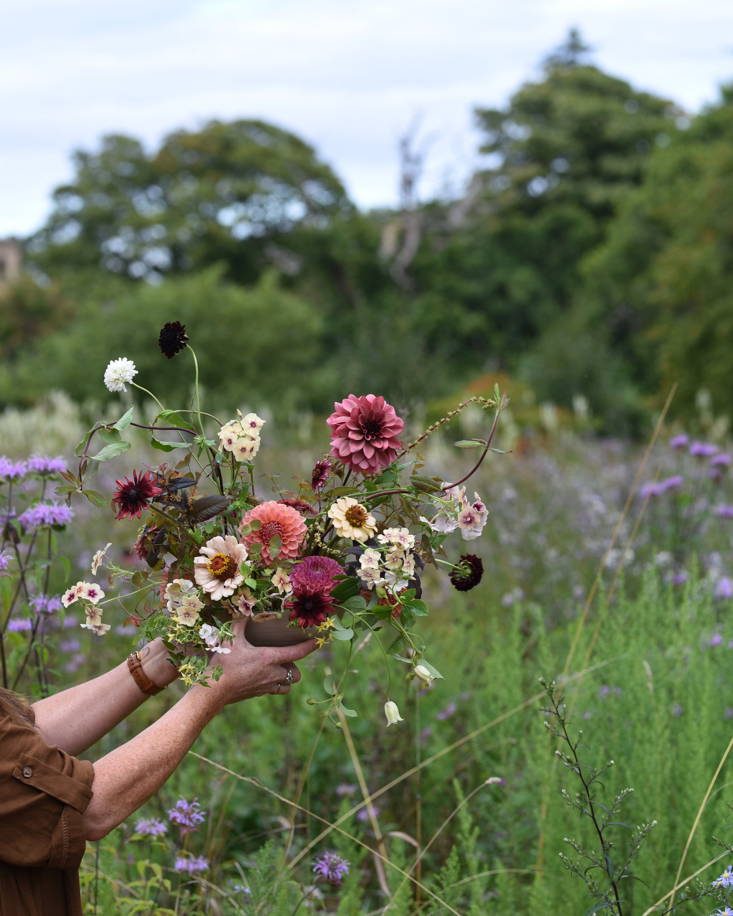 Person holding a colorful bouquet of flowers in a field of wildflowers, with trees and a cloudy sky in the background.