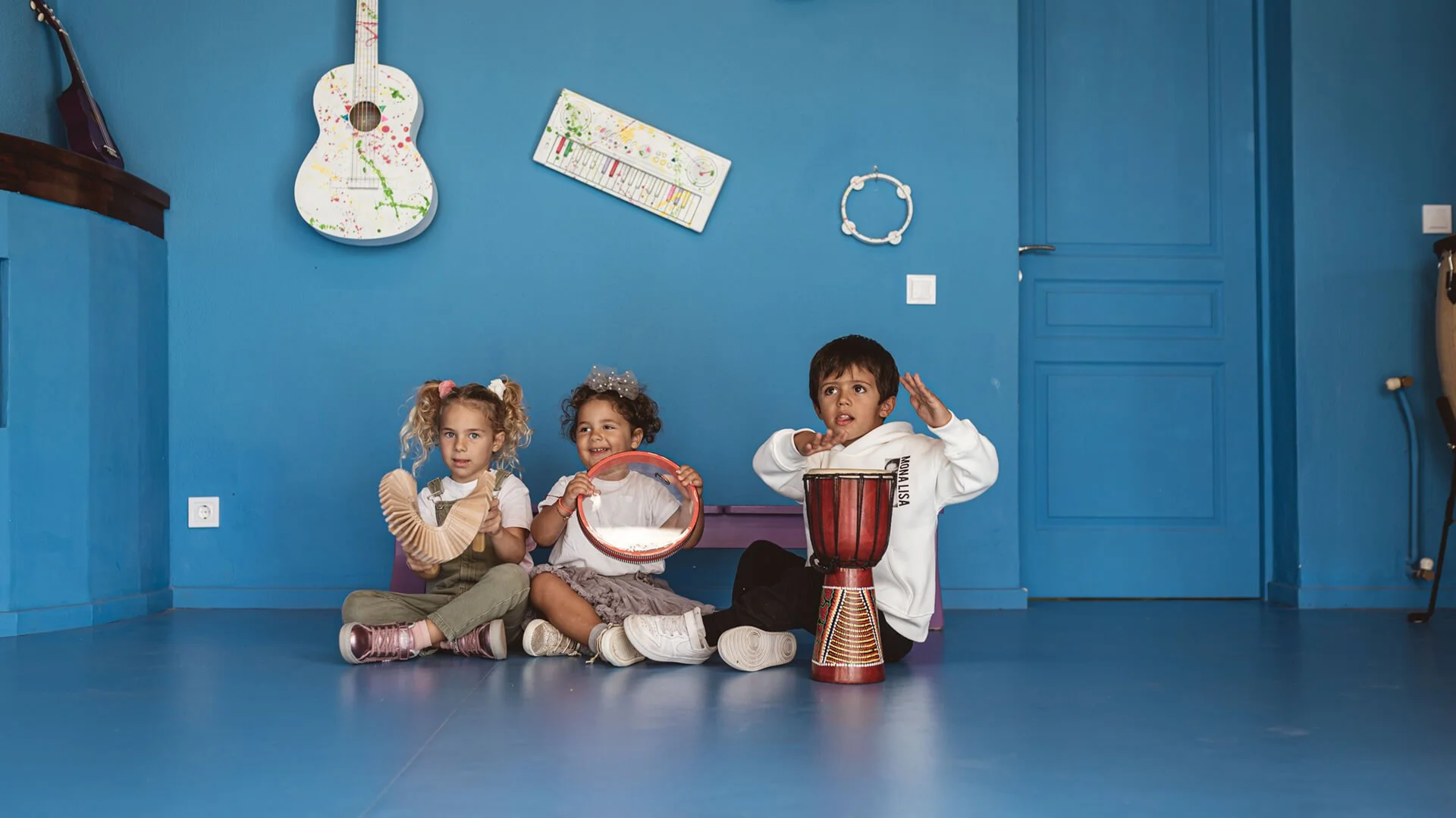 Three children sitting on the floor in a blue room, playing musical instruments. The girl on the left has curly blonde hair in pigtails and is holding a wooden instrument. The girl in the middle has curly hair with a bow and is holding a tambourine. The boy on the right has short dark hair, is dressed in a white hoodie, and is playing a drum. The room has blue walls with a painted white guitar, and a small whiteboard hanging on the wall.