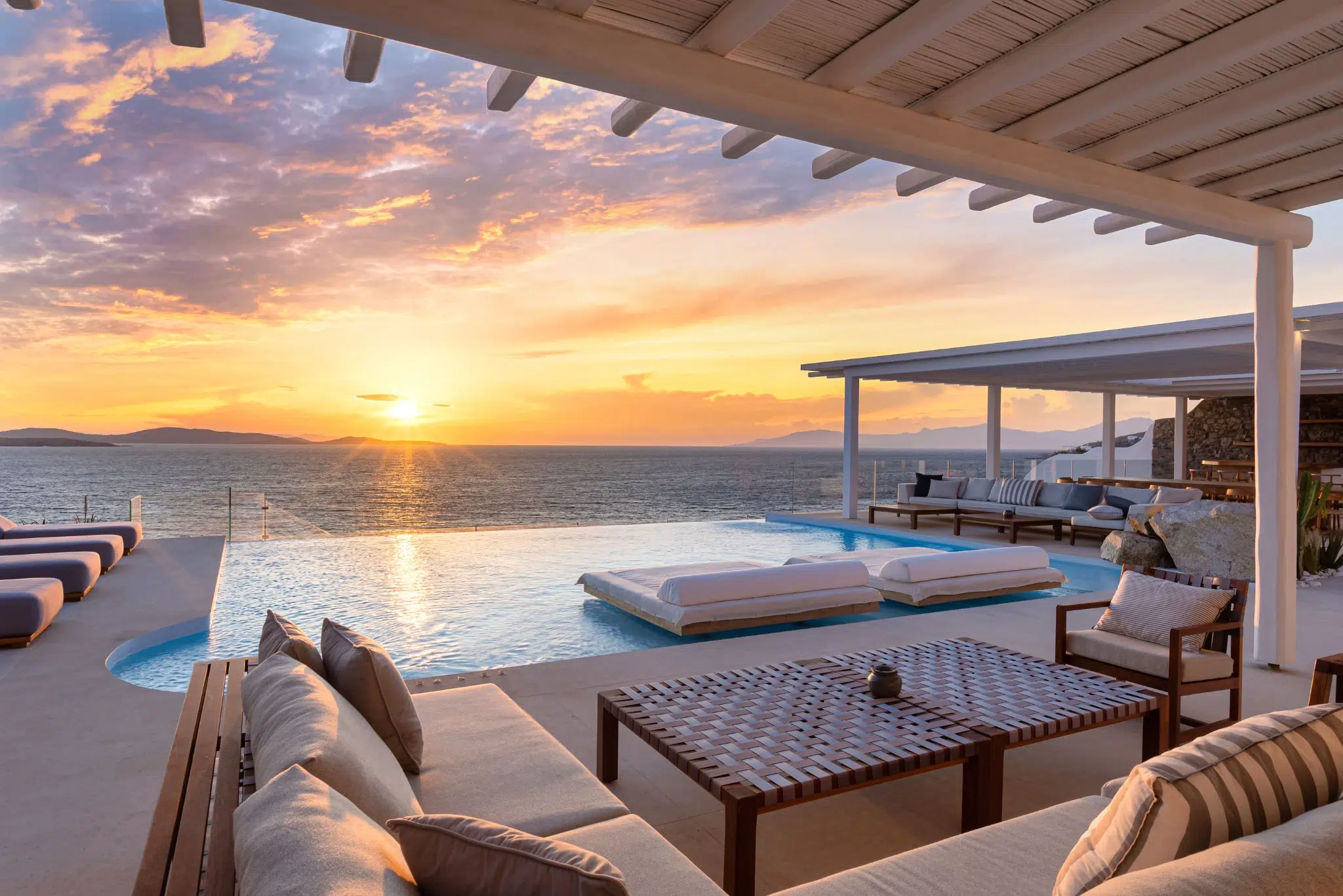 Outdoor lounge area overlooking ocean at sunset with pool, cushioned seating, and wooden furniture