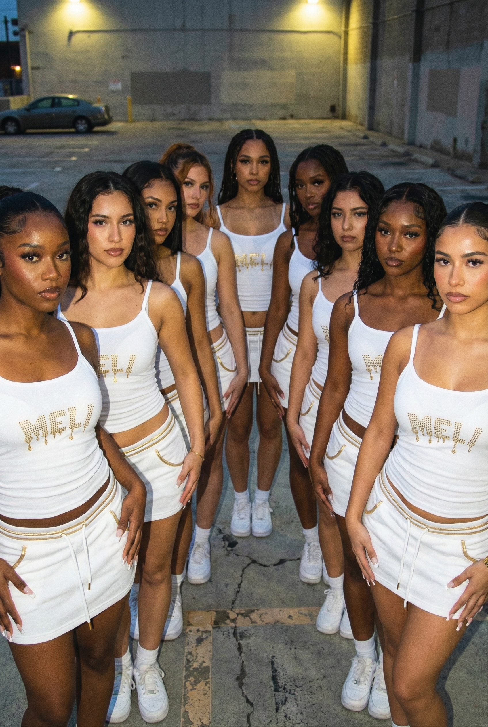 Group of women dressed in white athletic wear with gold accents, standing in a parking lot at night. Featuring white cropped cami tank top with rhinsetone Meli logo from Anastasia Giousef and Anais Meli Zoom Zoom sponsored by Deligiannis company