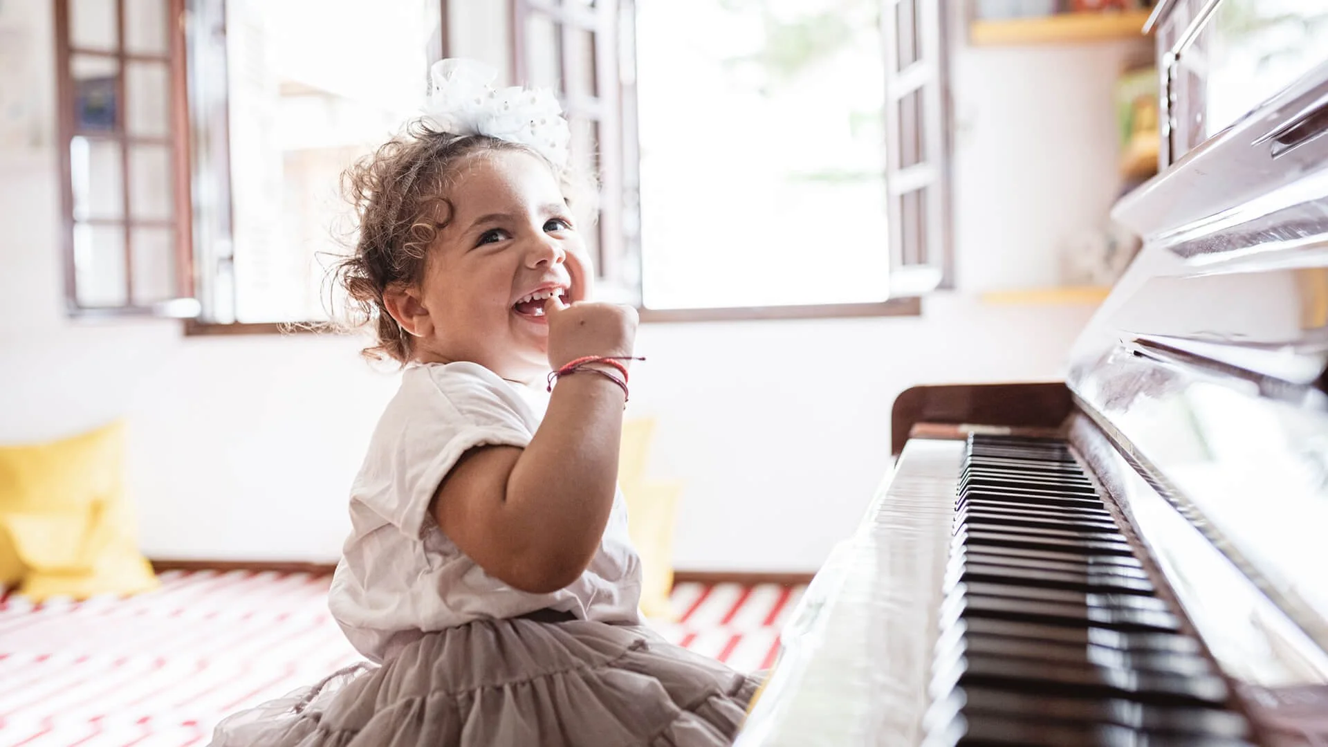 A young girl with curly hair, wearing a bow, sitting at a piano with a joyful expression, inside a bright room with windows and yellow decor.