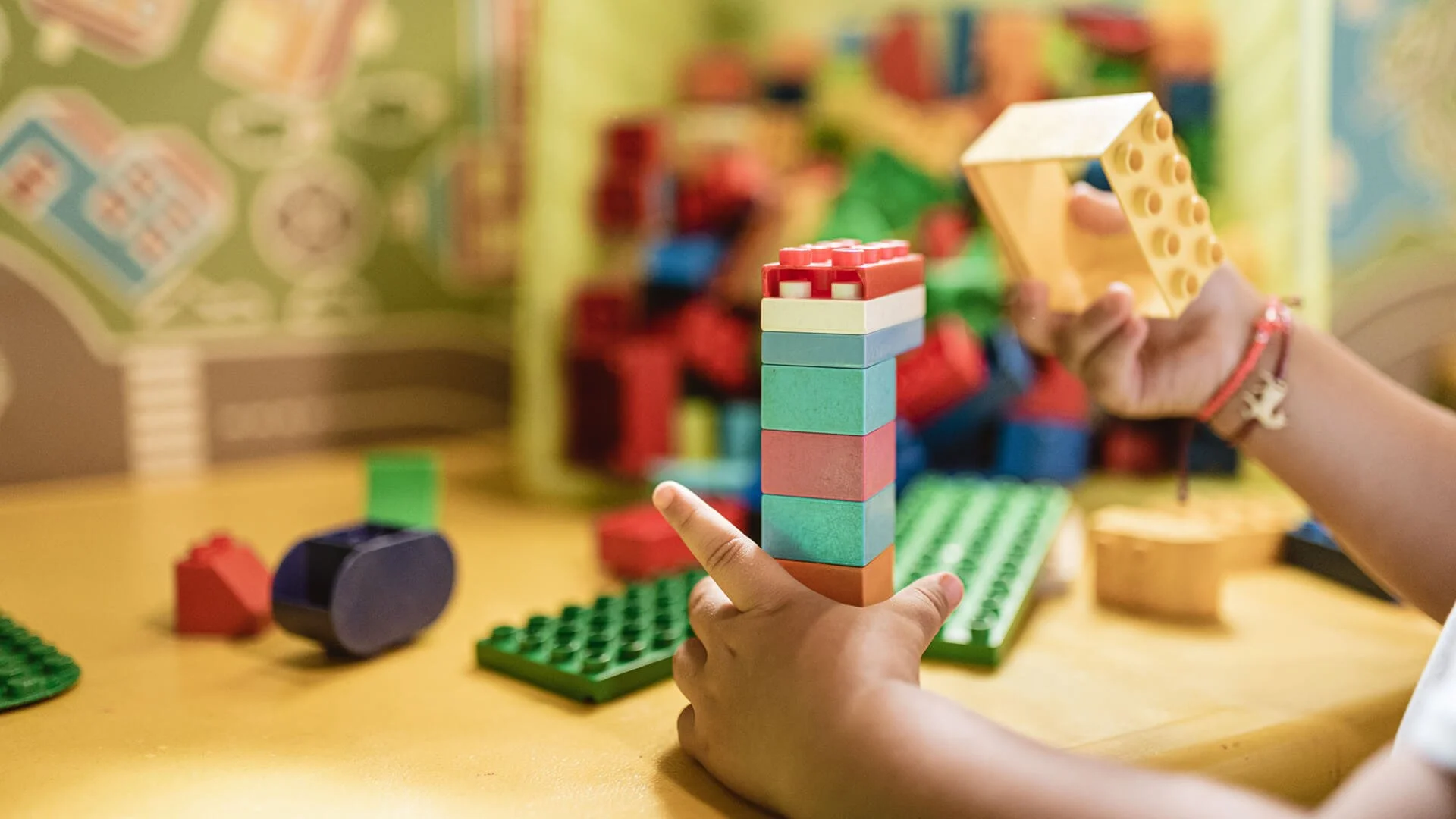 Child playing with colorful building blocks at a table with a green Lego baseplate and a poster with building instructions in the background.