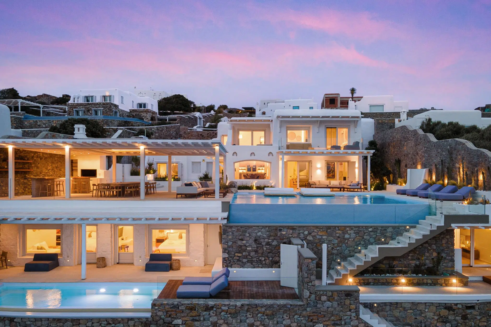Luxury white villa with multiple levels, outdoor swimming pool, patio furniture, and hillside background during sunset.