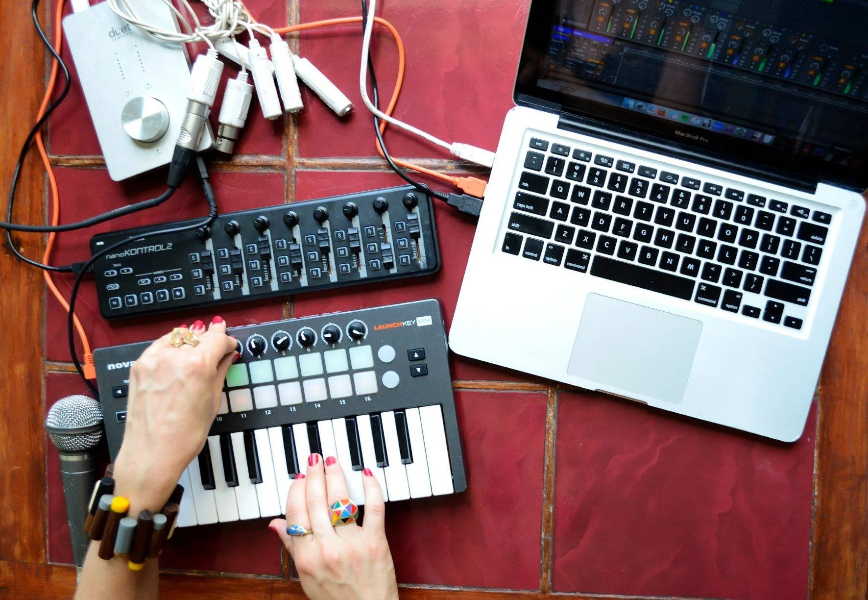 A laptop, a midi keyboard, a launchpad, a midi control surface, a microphone, and various audio cables on a red-tiled floor. A person with painted nails and multiple colorful rings is working on the launchpad.