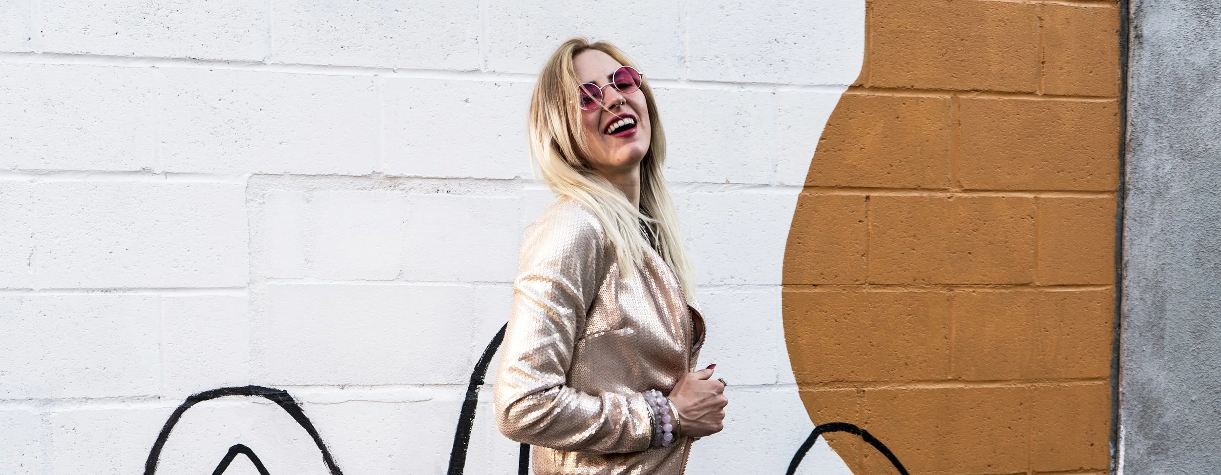 Woman with blonde hair wearing pink sunglasses, a shiny beige jacket, and smiling, standing against a white and orange mural wall. Alarke