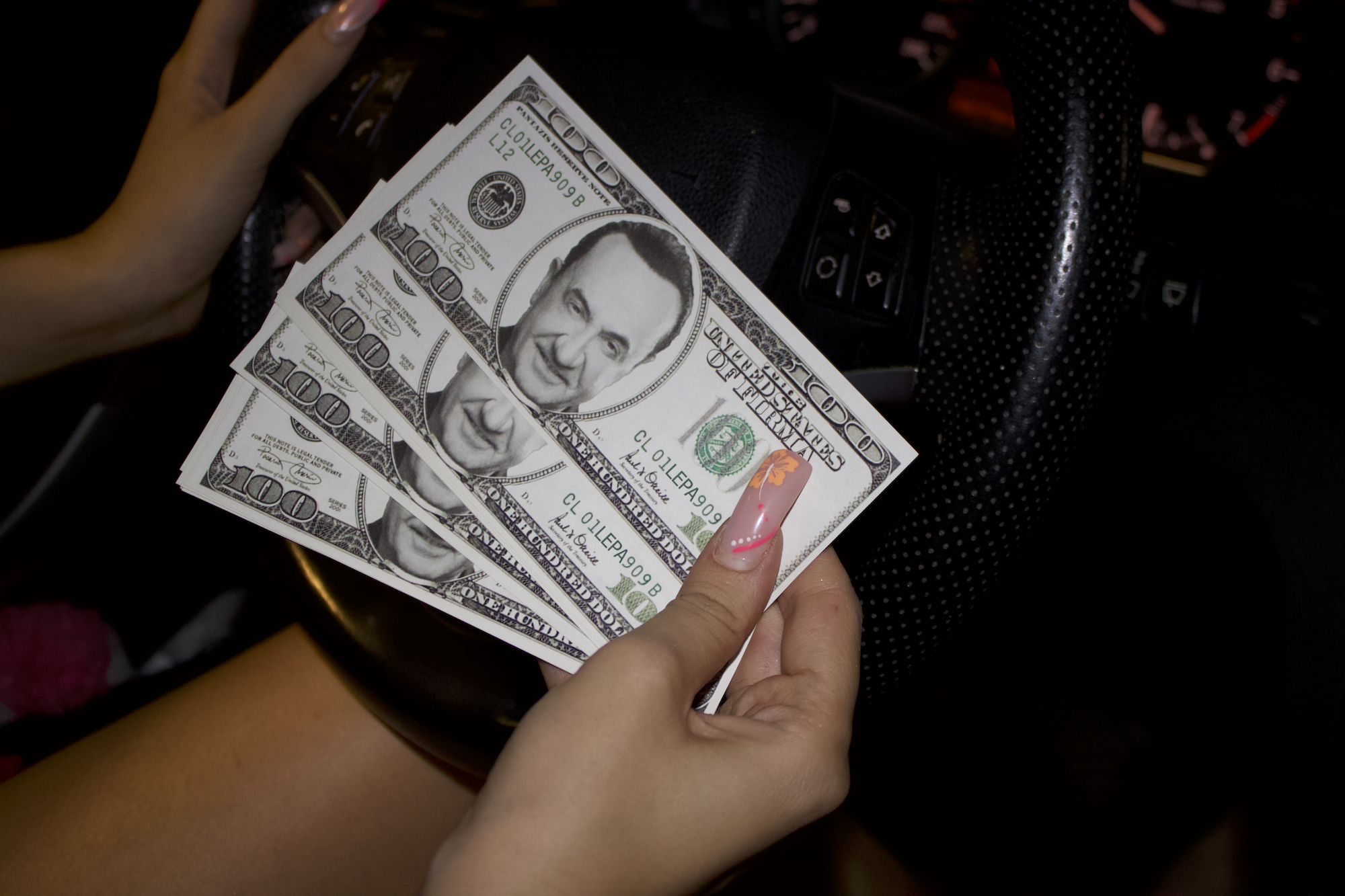 Person holding three $100 bills with the face of Lefteris Pantazis, Λευτέρης Πανταζής, ΛΕΠΑ, Lepa, inside a car, with a steered wheel and dashboard visible.