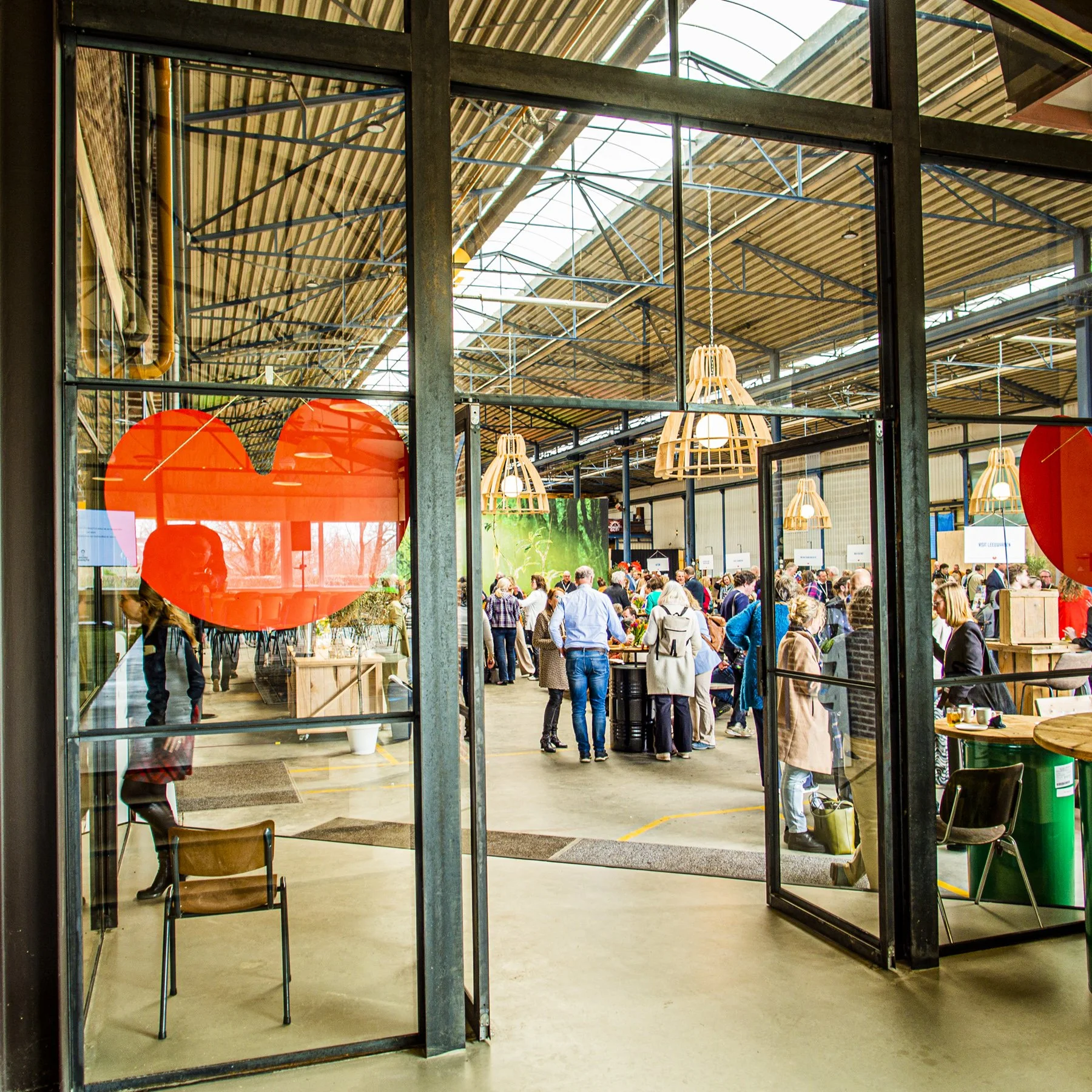 Binnenzicht van een drukke markt of evenement met mensen die rondlopen en aan tafels staan, Evenement lokatie Friesland. Hoge plafonds met houten en metalen structuren en hanglampen. Nijeleije vrij parkeren